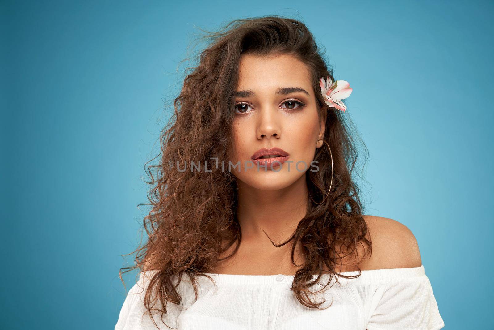 Front view portrait of attractive brunette woman with curly hair and in white sweater on blue background. Concept of modern shooting on professional photo studio. 