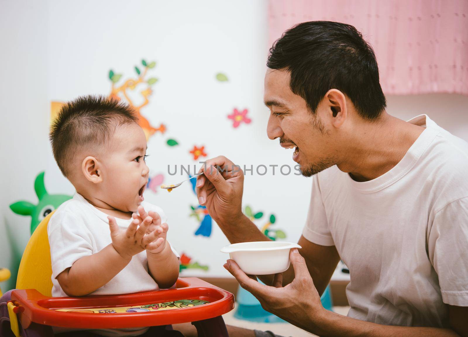 Father acting Mom feeding his son baby 1 year old on chair in the house