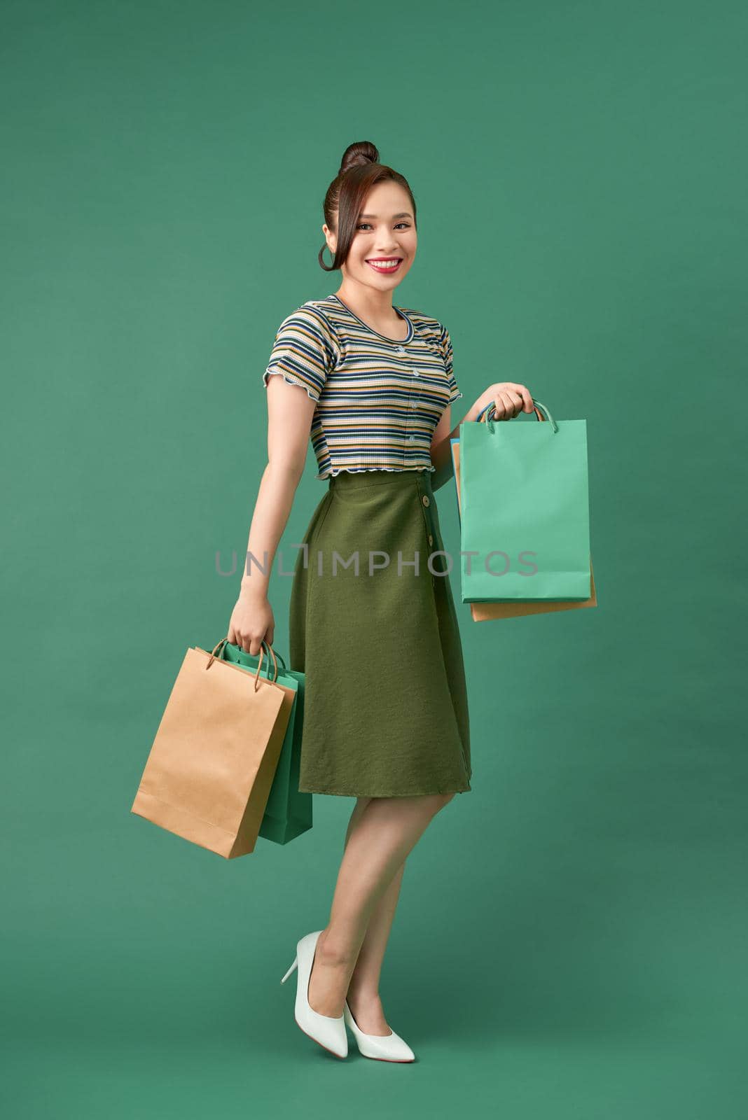 full length view of cheerful, fashionable woman holding shopping bags and smiling on green background