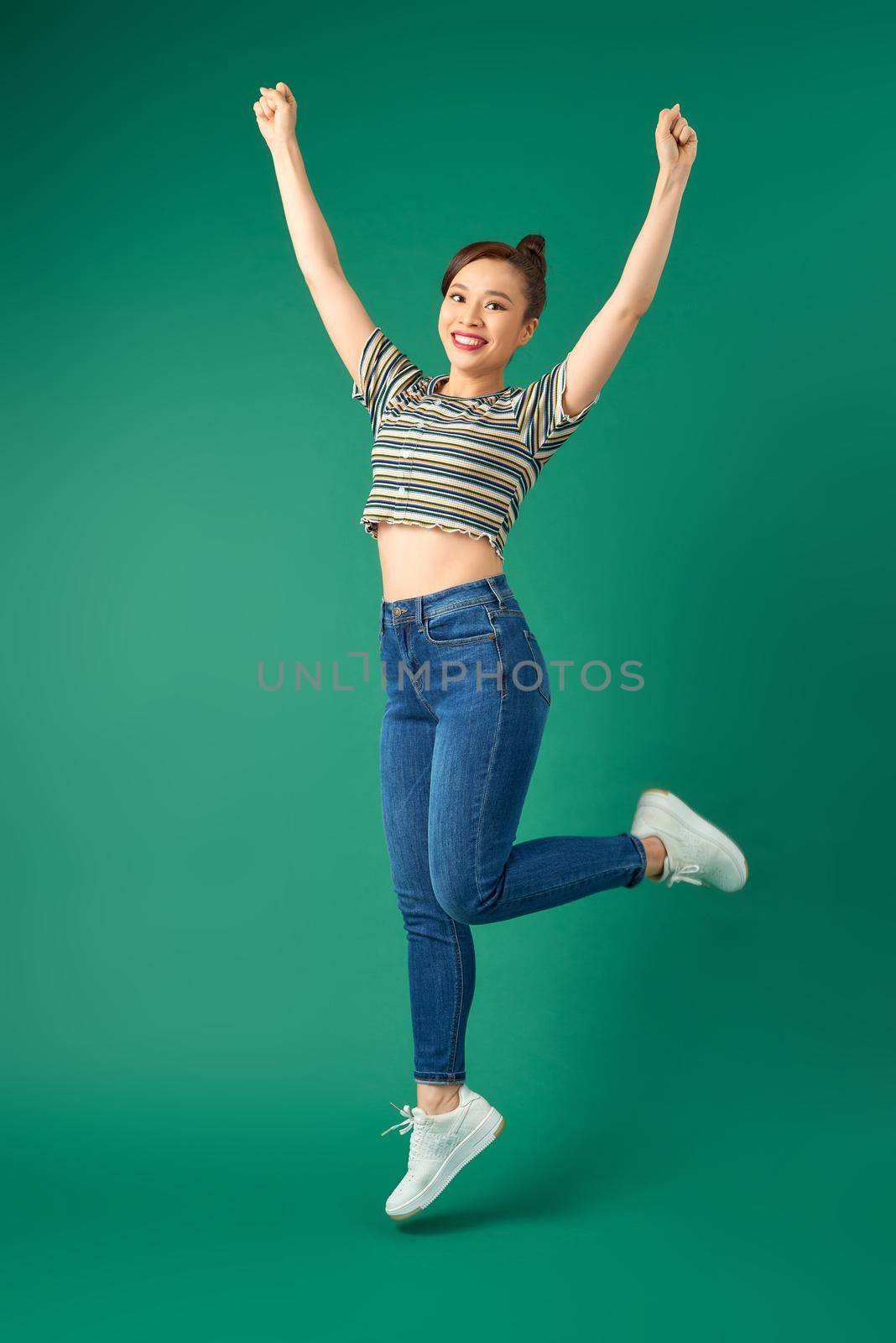 Full-length portrait of cheerful positive young Asian woman jumping over green background.