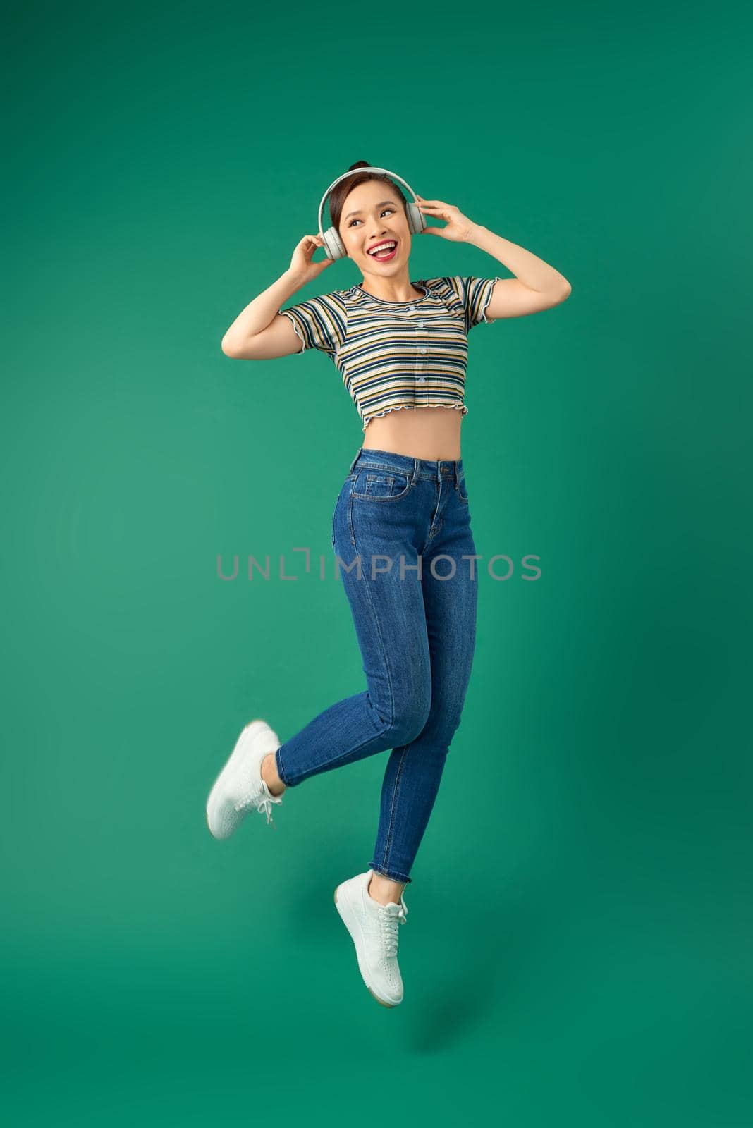 Full length portrait of young Asian woman in headphones listening to music dancing isolated green background