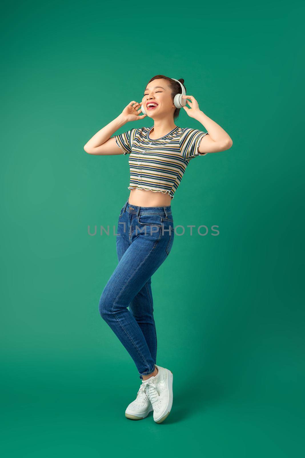 Energy girl with headphones listening to music with closed eyes on green background in studio. Wearing T-shirt, jeans.