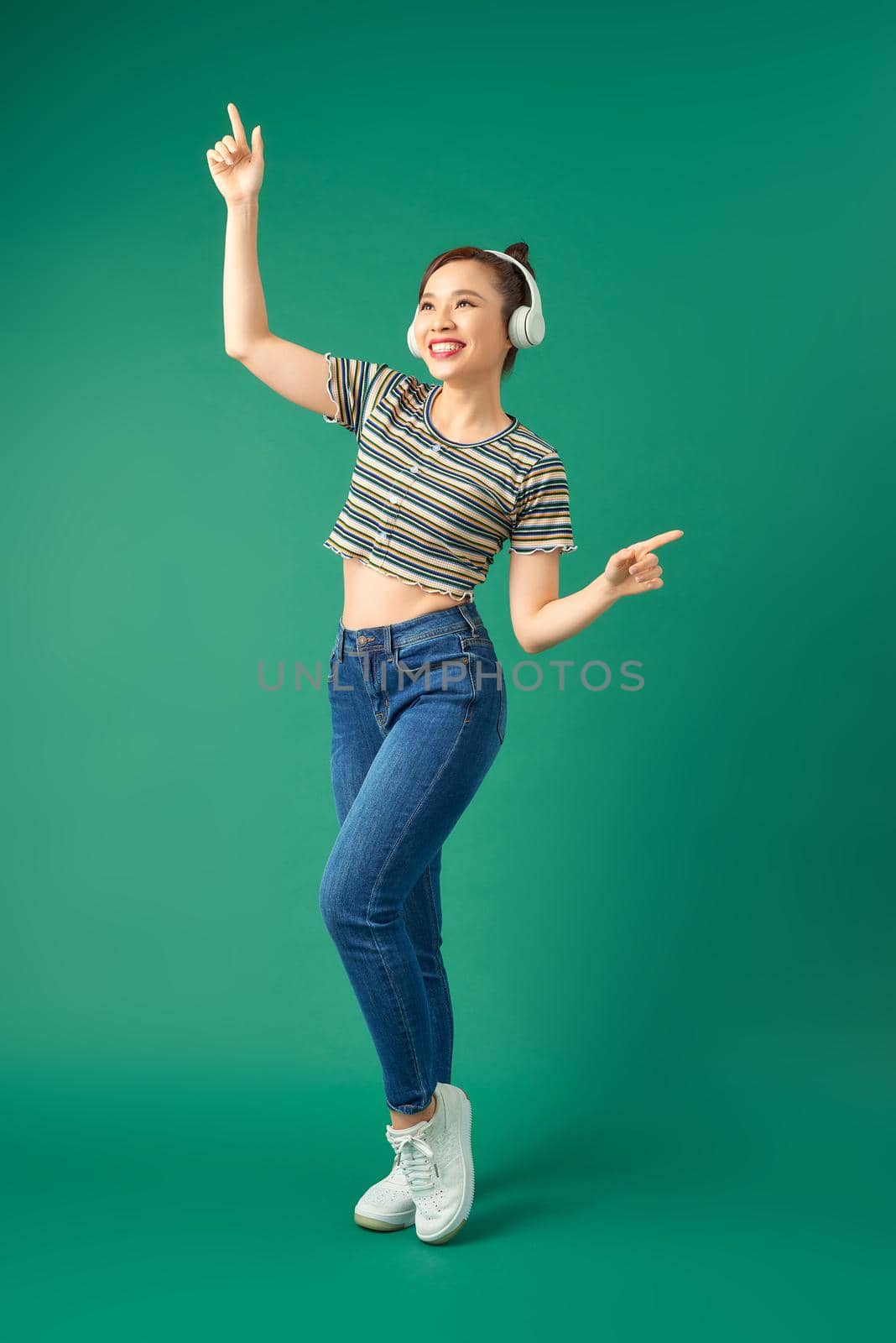 Full length portrait of joyful Asian woman on casual clothing dancing and listening to music with headphone over green background.