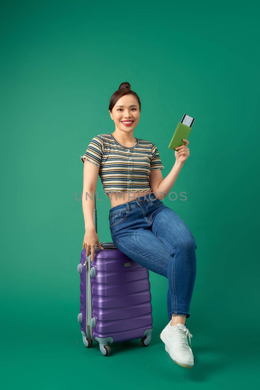 Attractive young Asian woman sitting on suitcase and holding passport, ticket flight over green background. by makidotvn