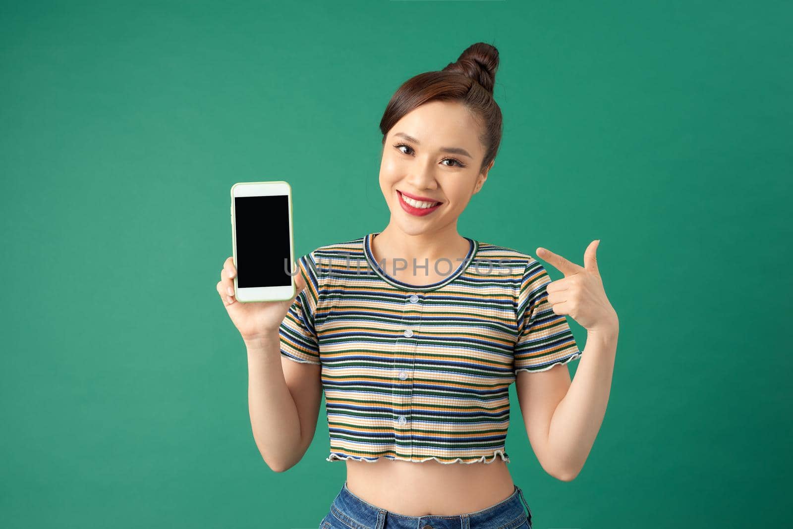 Smiling young Asian woman pointing with index finger at blank screen phone isolated over green background.