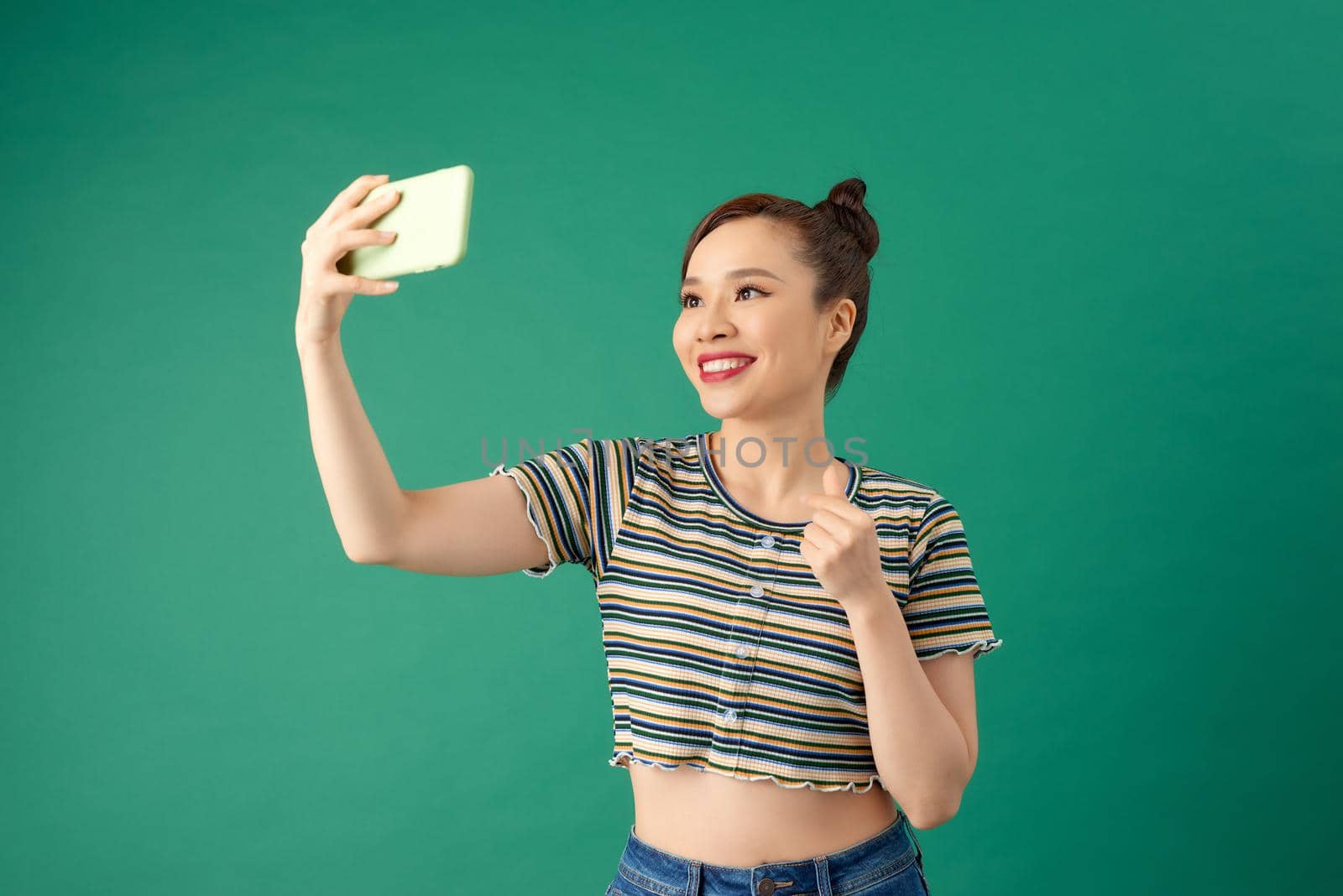 Portrait of young Asian female making selfie photo on smartphone with postitive expression over green background.