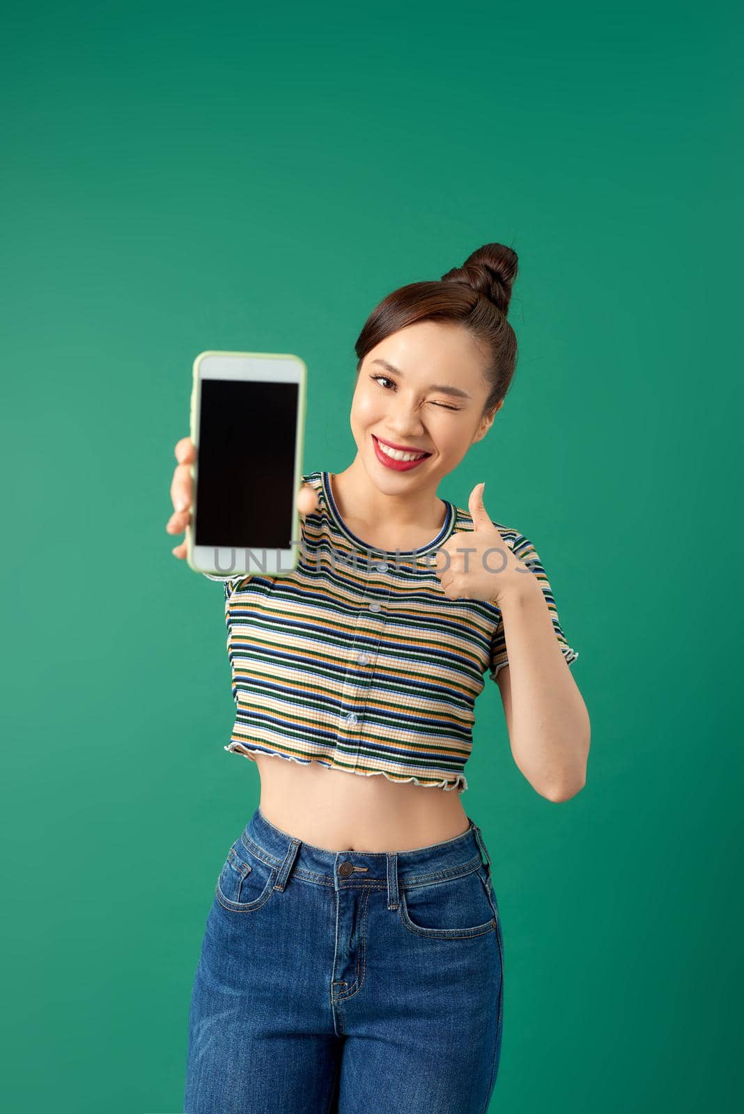 Young beautiful Asian woman in casual style showing blank screen mobile phone while standing over green background.