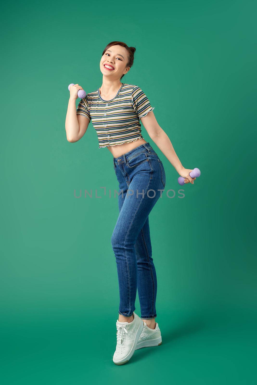 Attractive young Asian woman holding dumbell while standing over green background. Full length.
