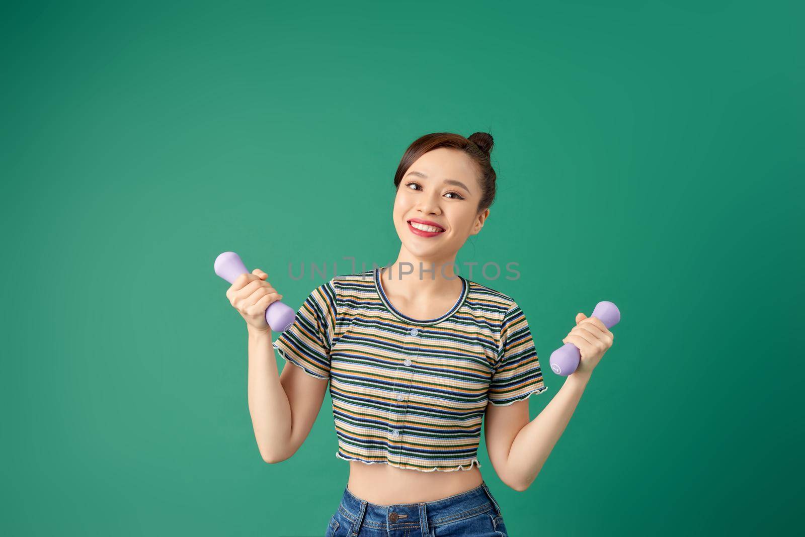 Smiling attractive young Asian woman practicing exercise with dumbell over green background.