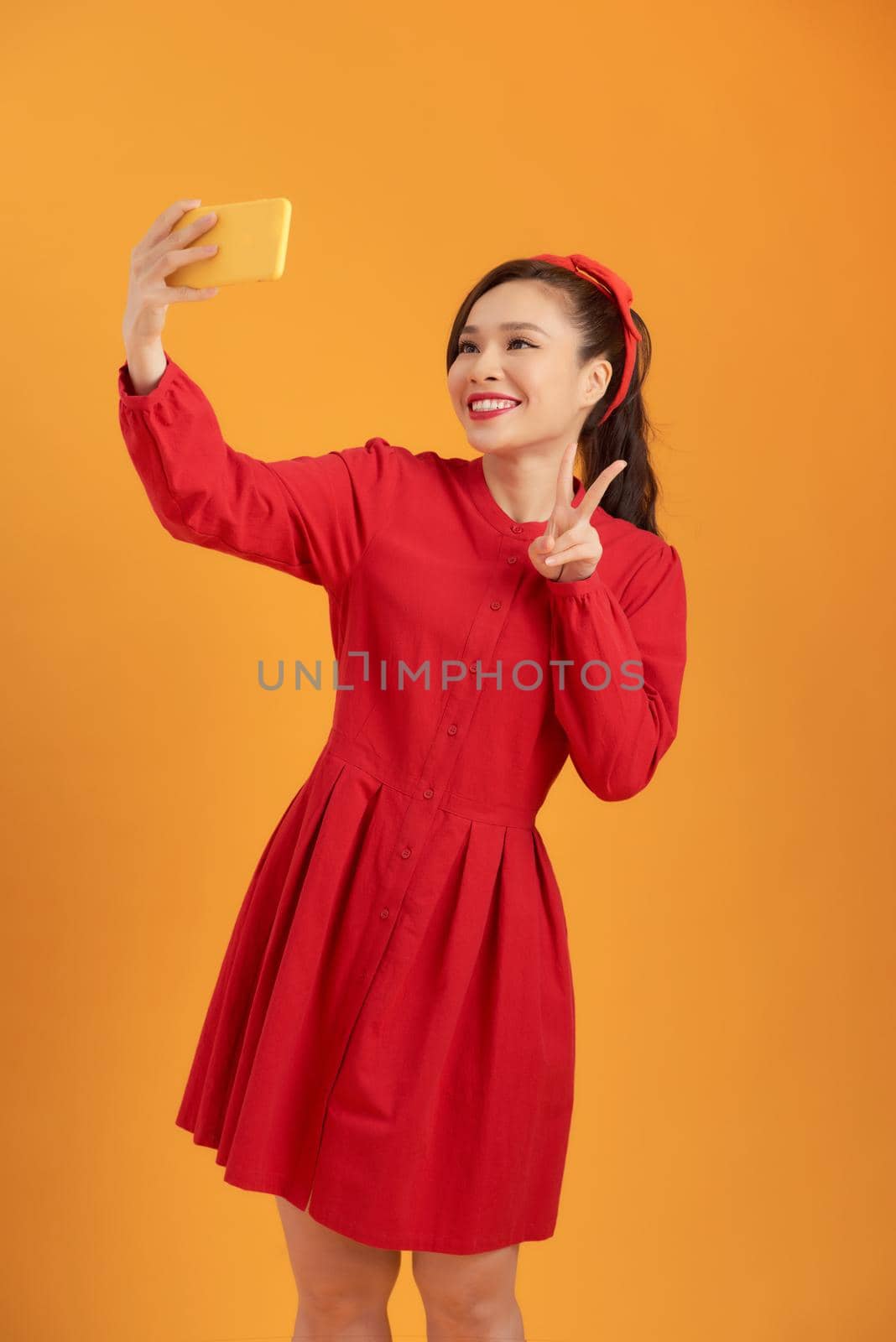 Shot of a happy young Asia woman taking selfie with her cellphone while standing isolated over orange background. by makidotvn