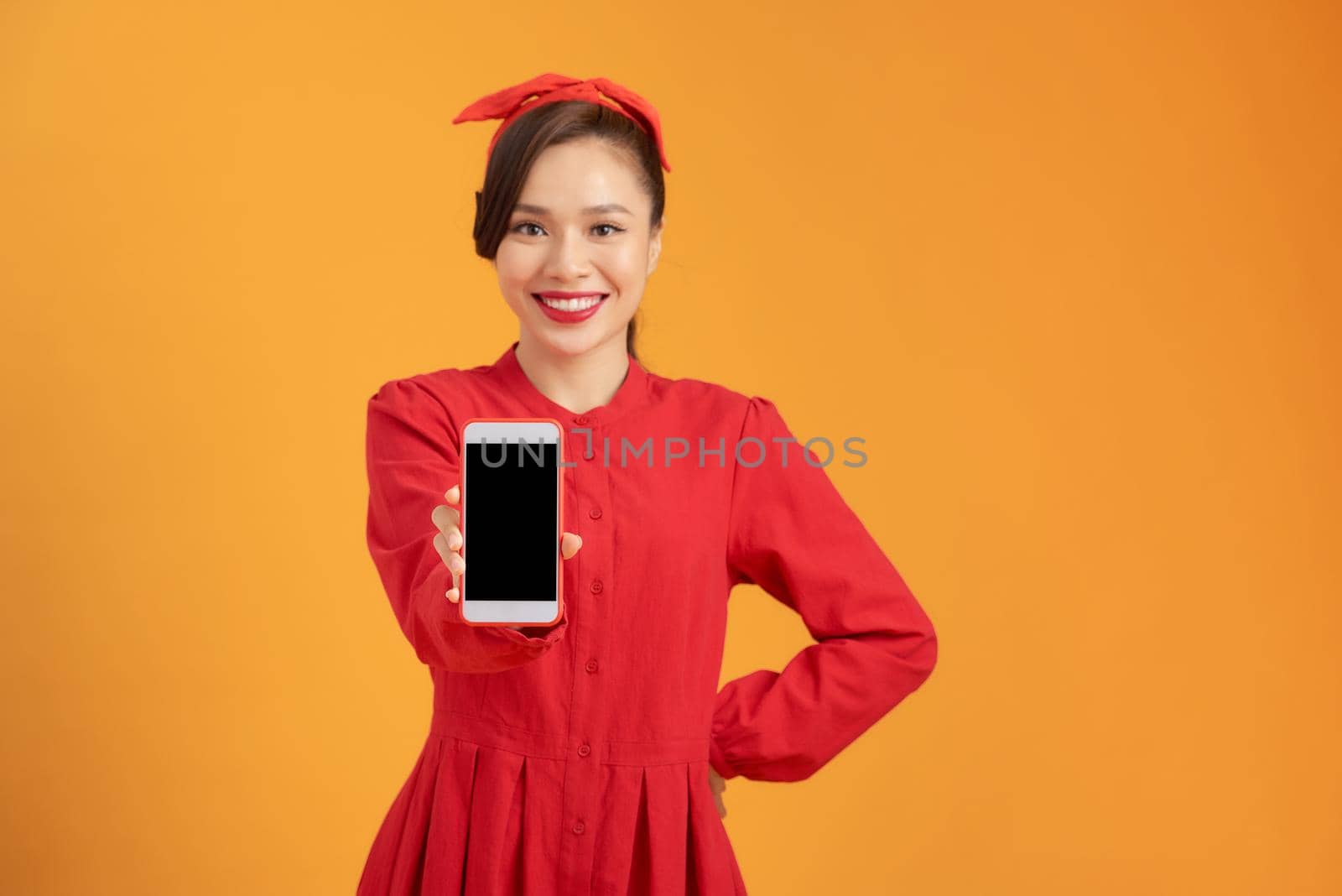 Portrait of a smiling asian woman showing blank screen mobile phone while standing isolated over orange background