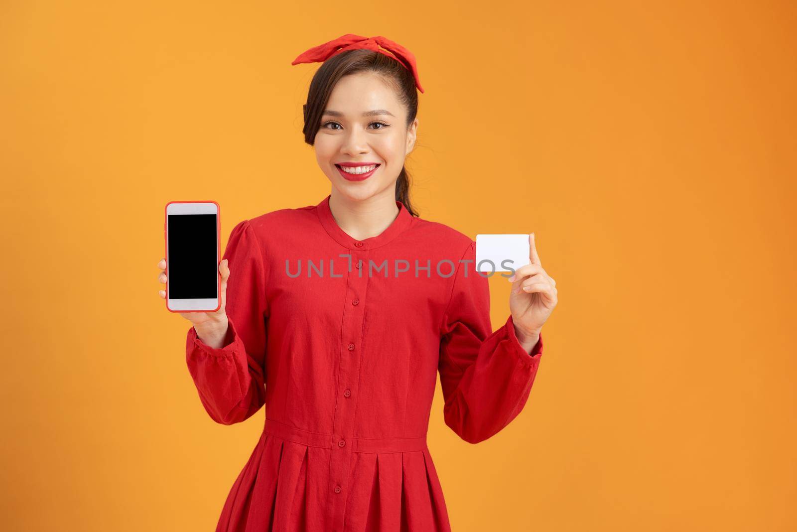 happy young Asian woman shows blank credit card and smart phone over orange background.