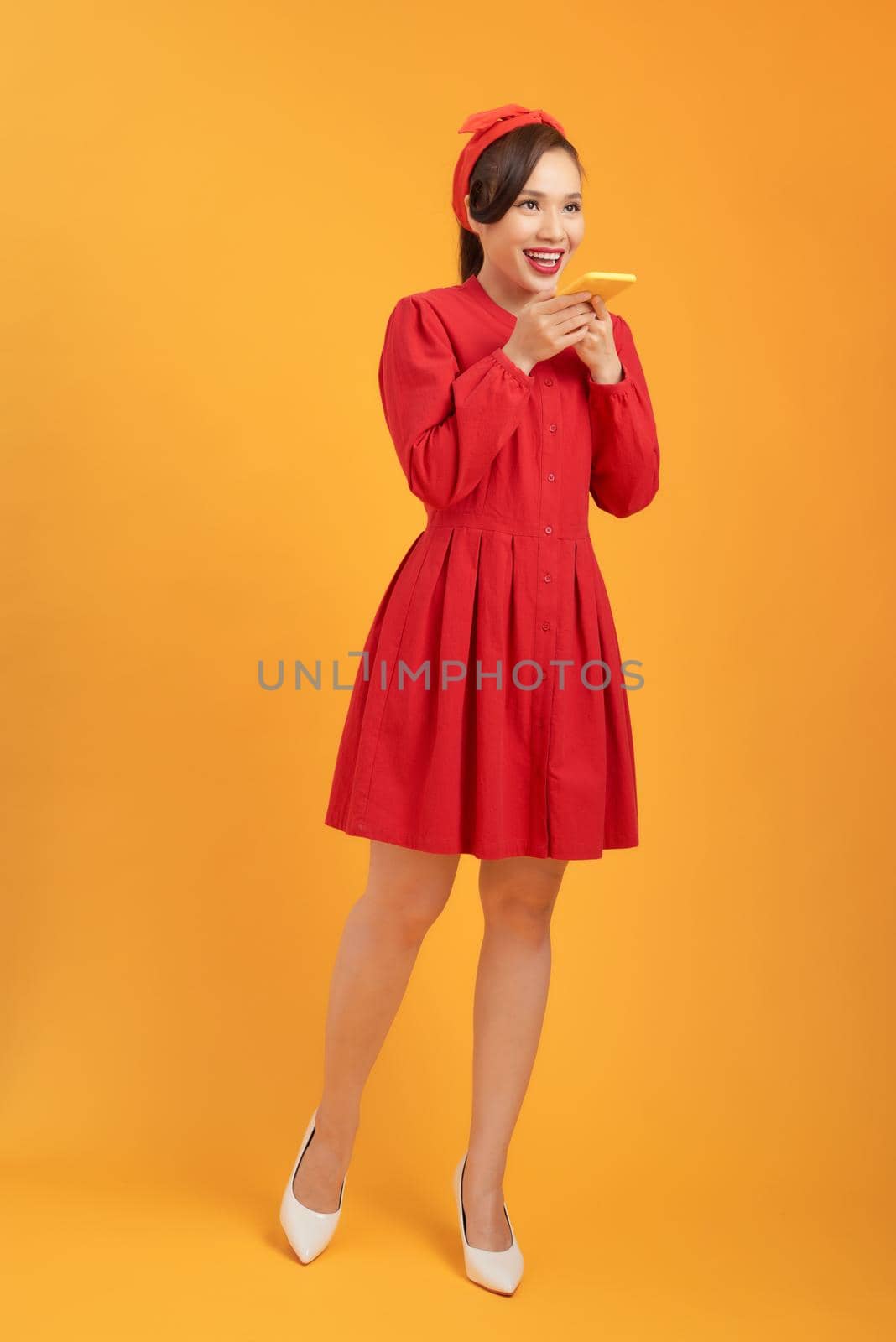 Full length portrait of a happy asian woman using mobile phone isolated over orange background.