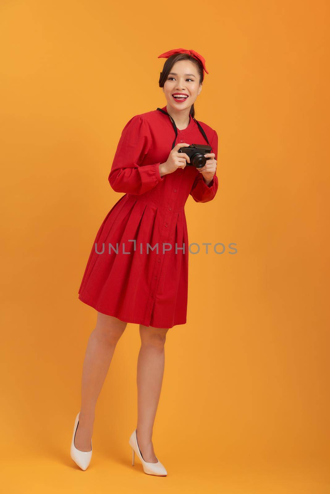 Smiling young Asian girl holding photo camera isolated on a orange background