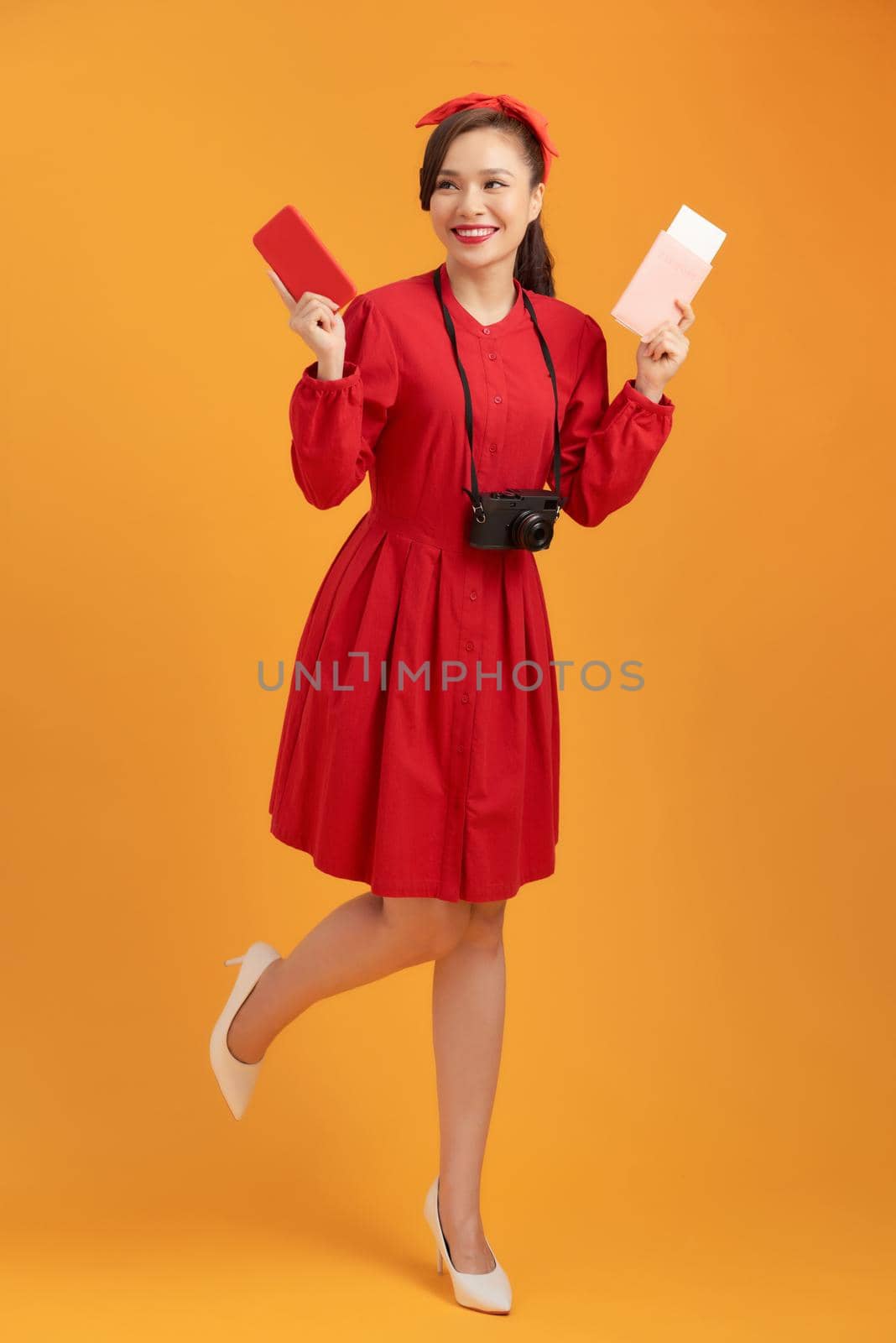 Happy young Asian woman tourist holding phone, passpoer, camera and standing over orange background. by makidotvn