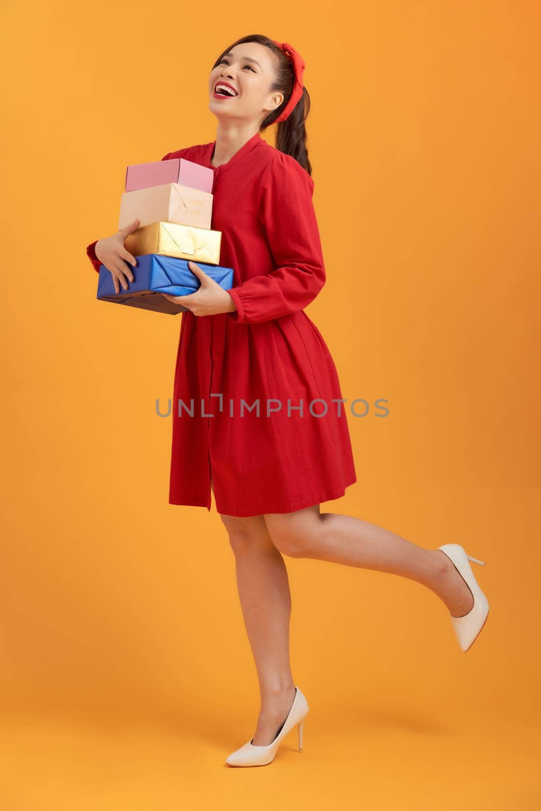 Happy young Asian woman holding stock of gift boxes over orange background.