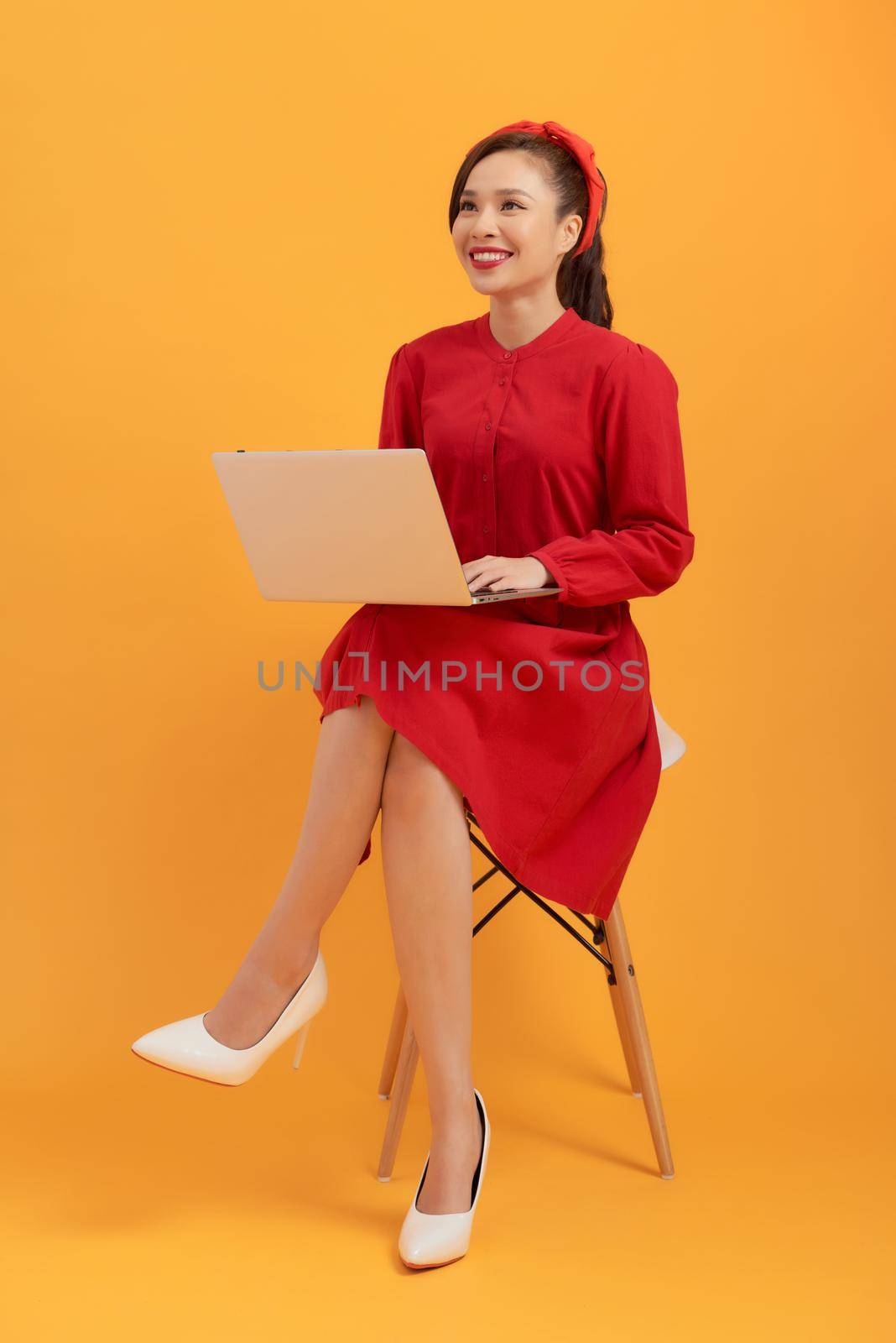 Beautiful young Asian woman using laptop and sitting in armchair isolated over orange background. by makidotvn