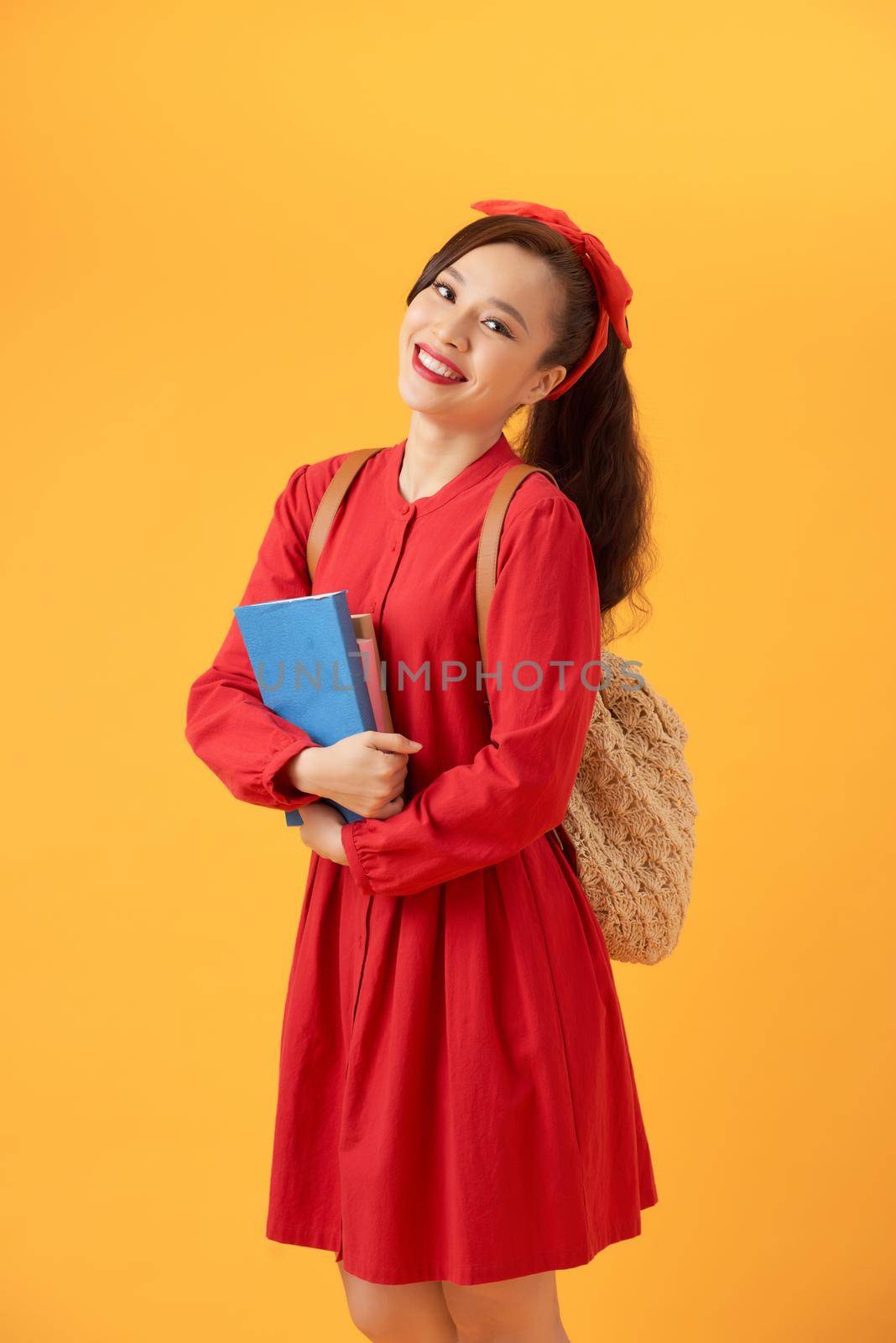 Beautiful Asian student holding books and bringing bag over orange background.