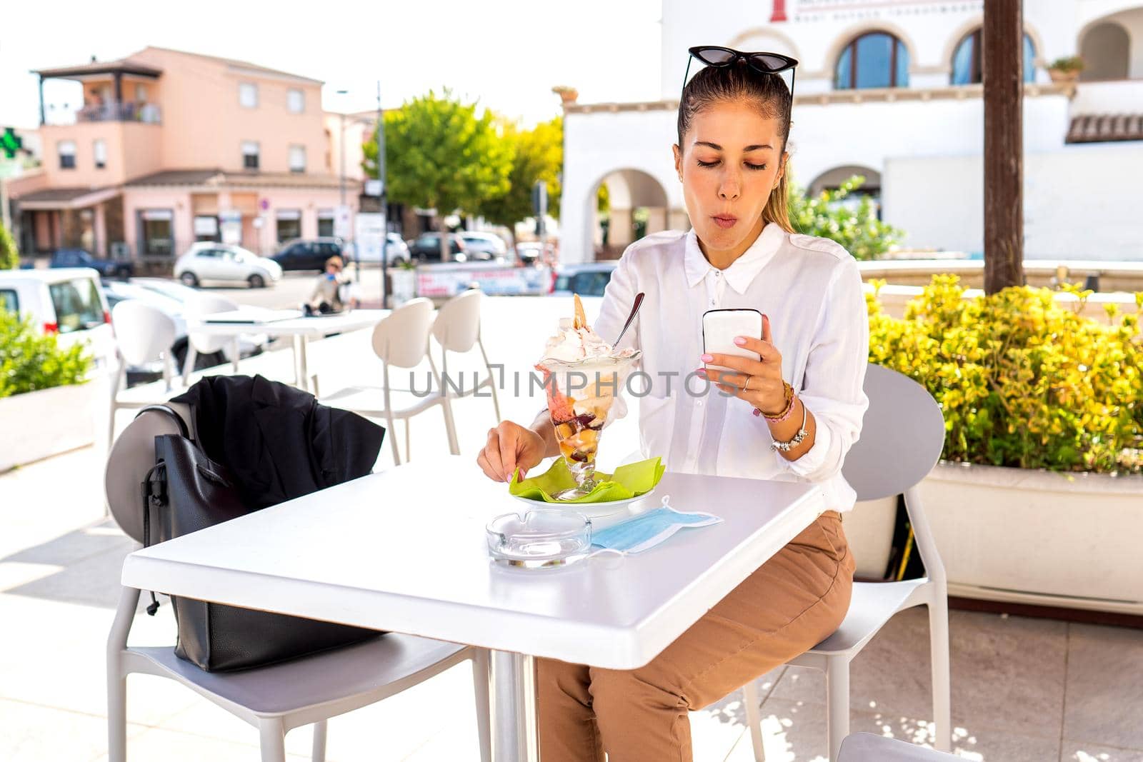 Beautiful modern elegant woman sitting at a dehore cafe bar table enjoying her big fruit flavored ice cream using mobile phone with internet connection to check email and chatting on social network