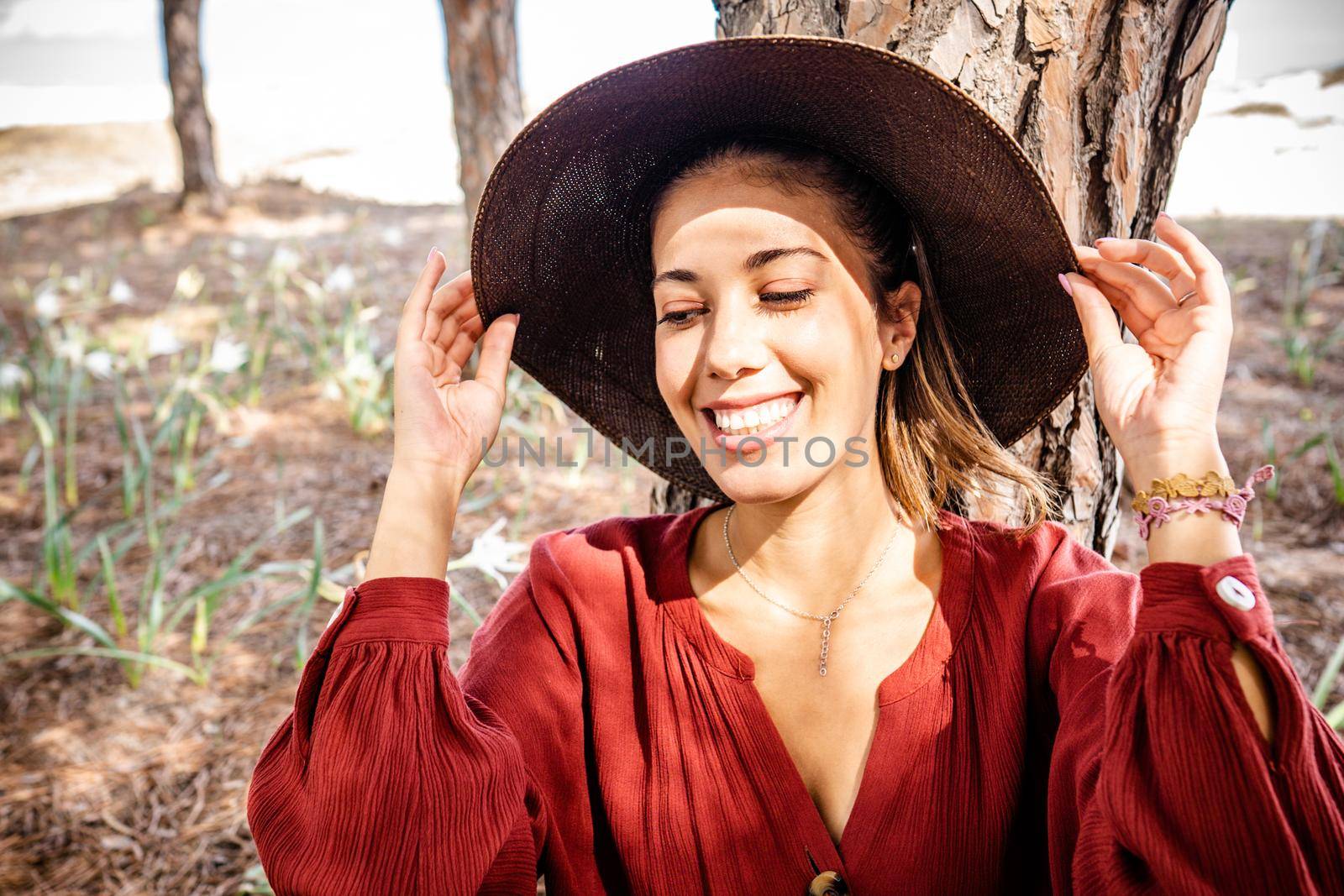Toothy smiling Beautiful candid young model , wearing red shirt standing in nature pine forest illuminated from the sun holding with her hands brims of her wide hat. Fashion shoot for spring concept