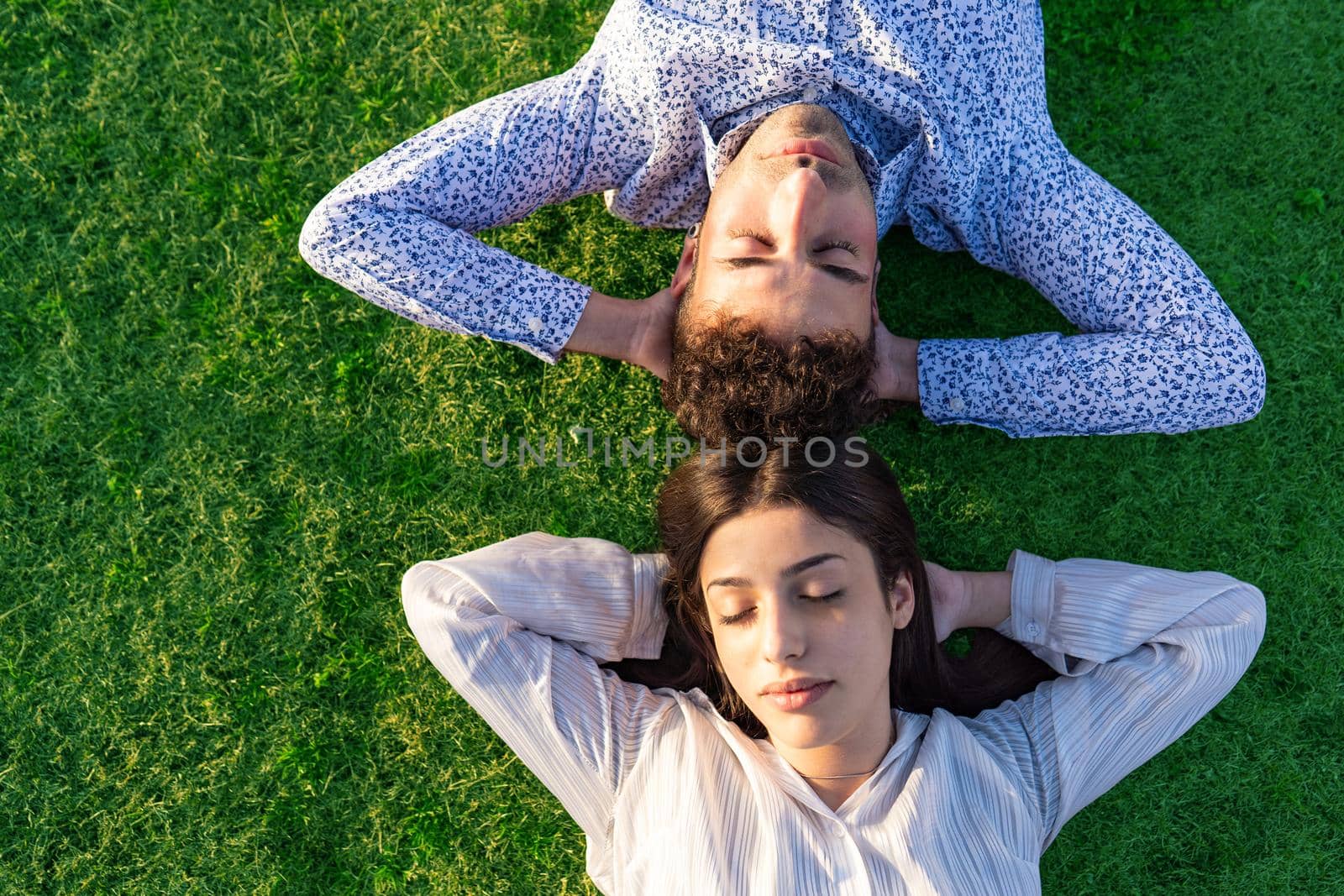 Top view of carefree young heterosexual couple resting sleeping on a beautiful green lawn with hands under heads. Two people relaxing enjoying together living the nature to healthy and better life