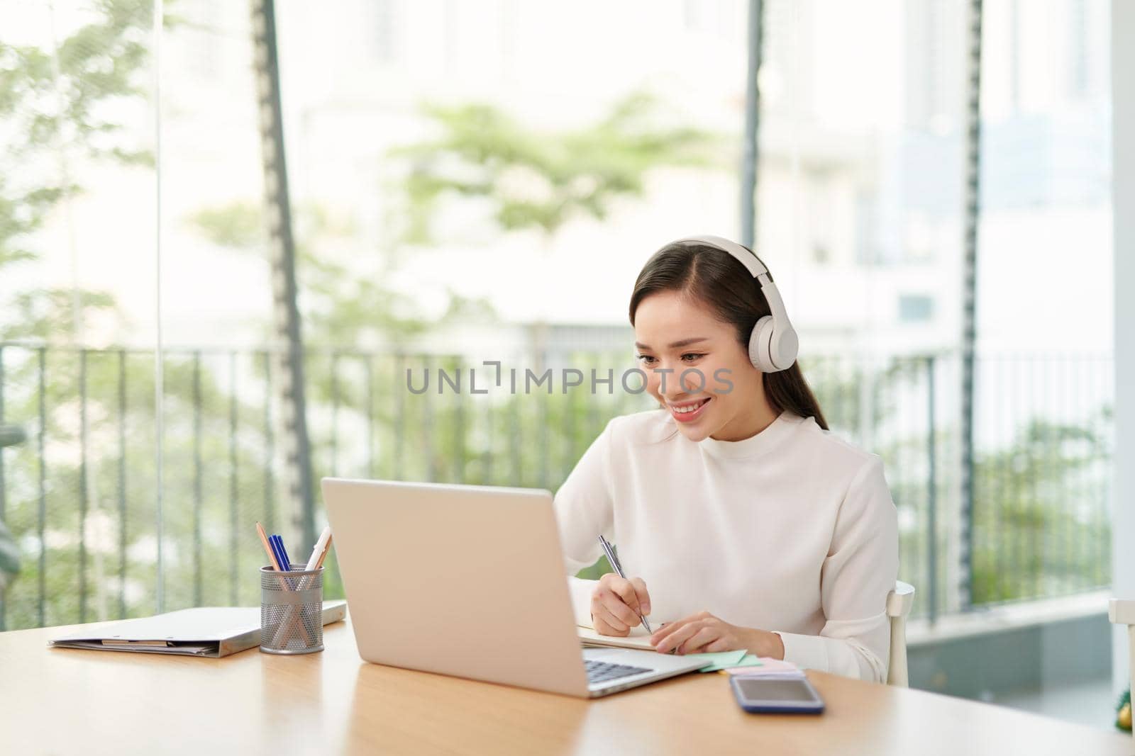 Smiling young woman sitting at the kitchen, using laptop computer for study, listening to music with headphones