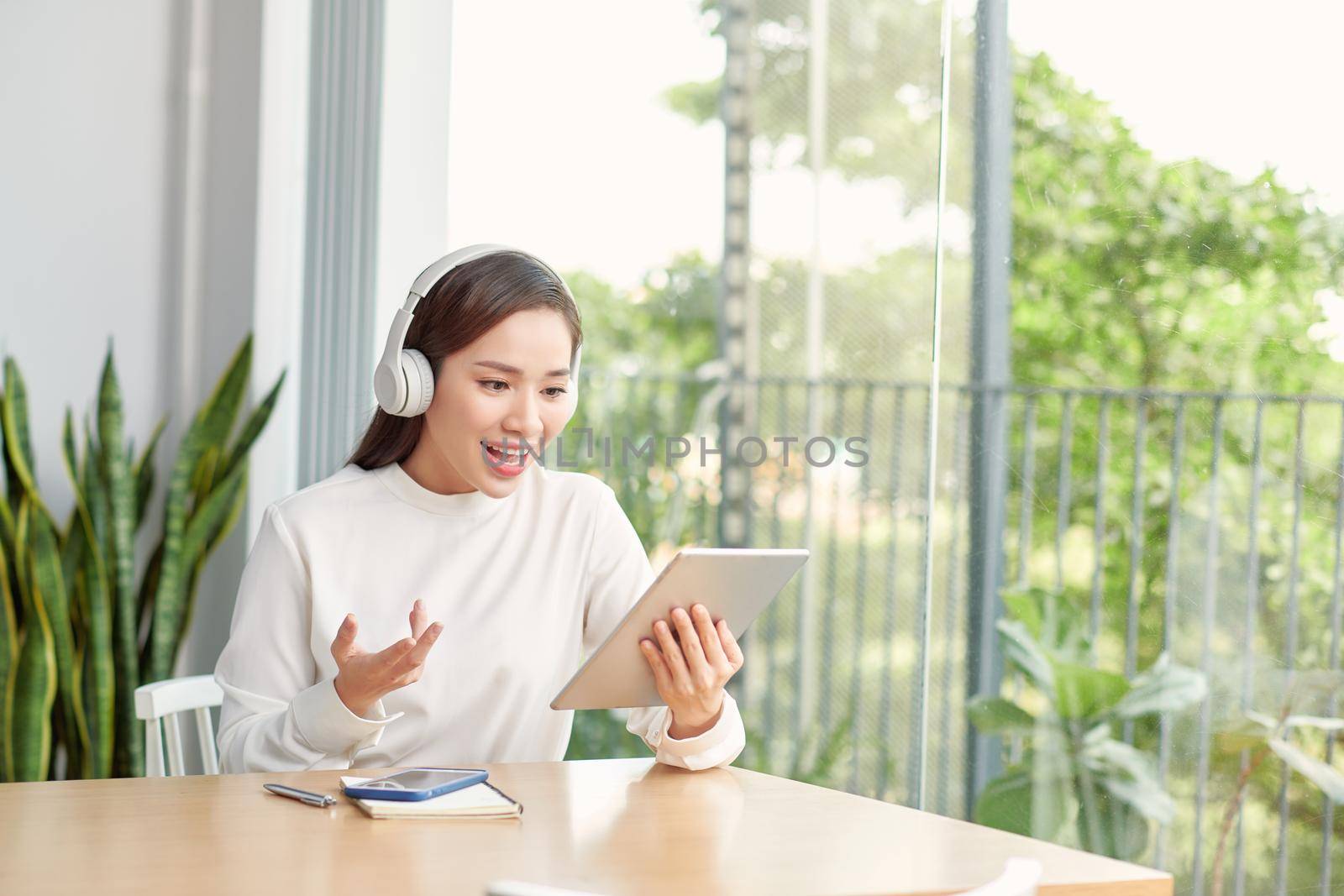Happy woman wearing wireless headphones waving in video conference on tablet sitting in hall of university