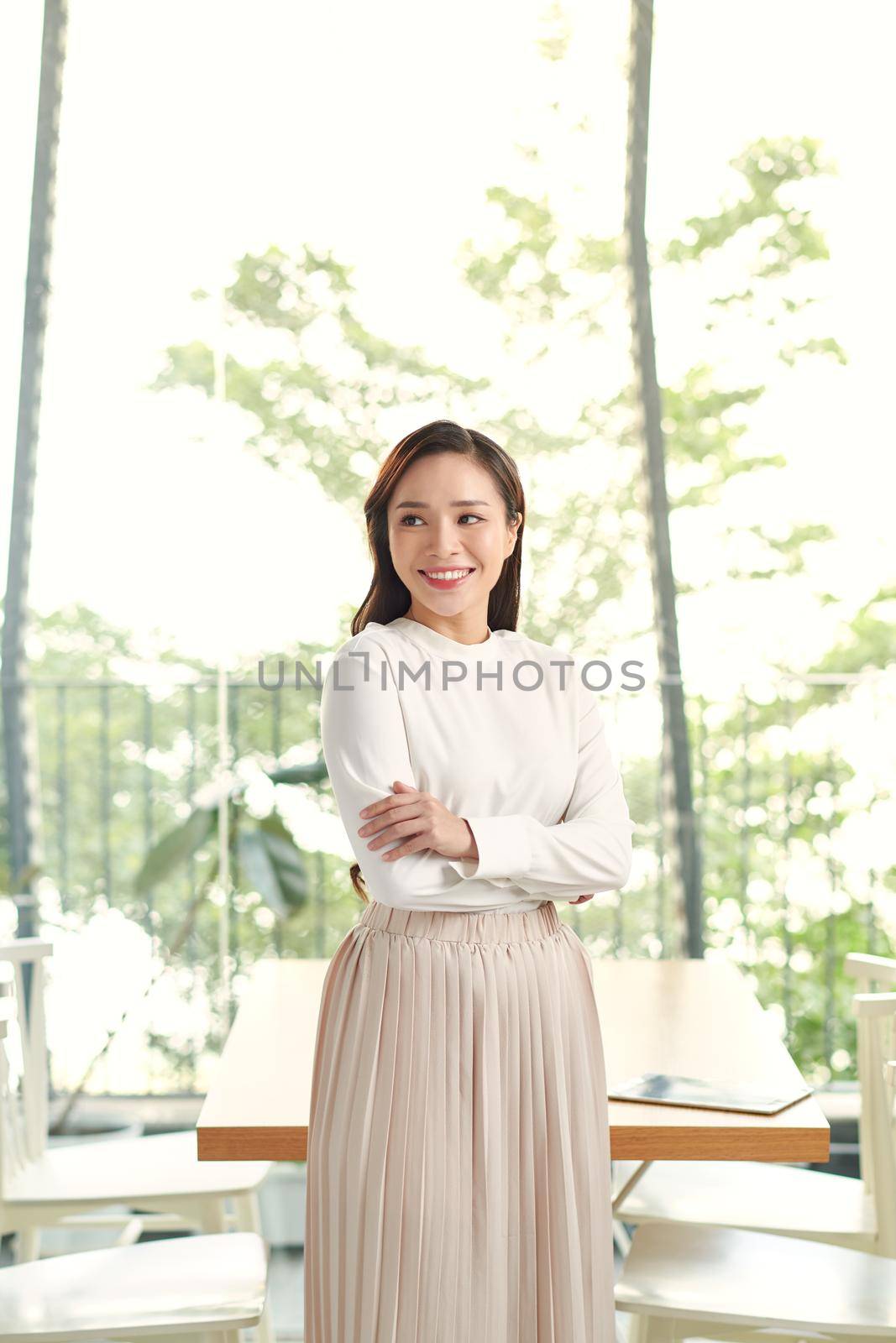 Happy young beautiful Asian woman with brace retainers, standing arms crossed, smiling to camera in modern office with big glass window and blur green background by makidotvn