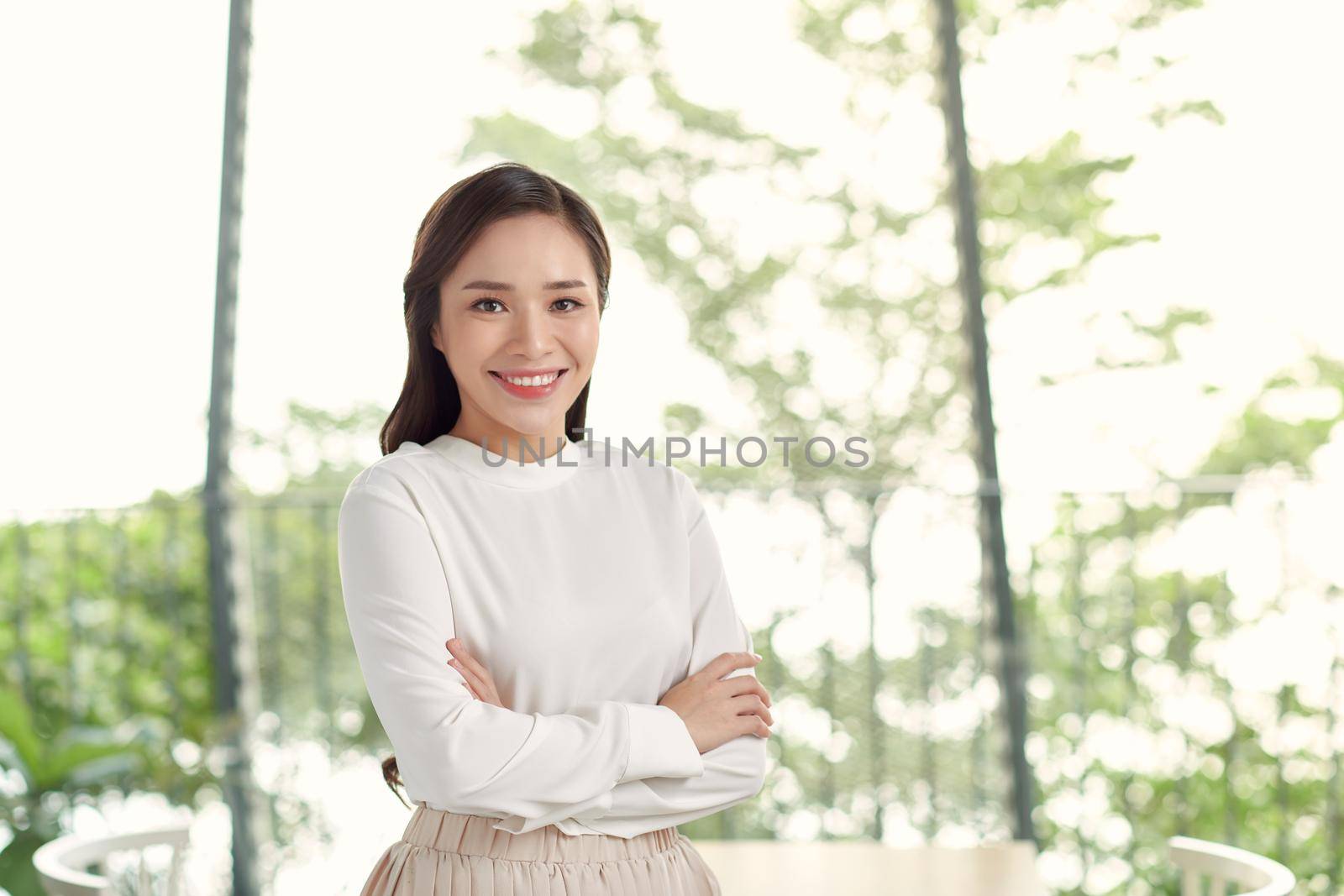 Happy young beautiful Asian woman with brace retainers, standing arms crossed, smiling to camera in modern office with big glass window and blur green background