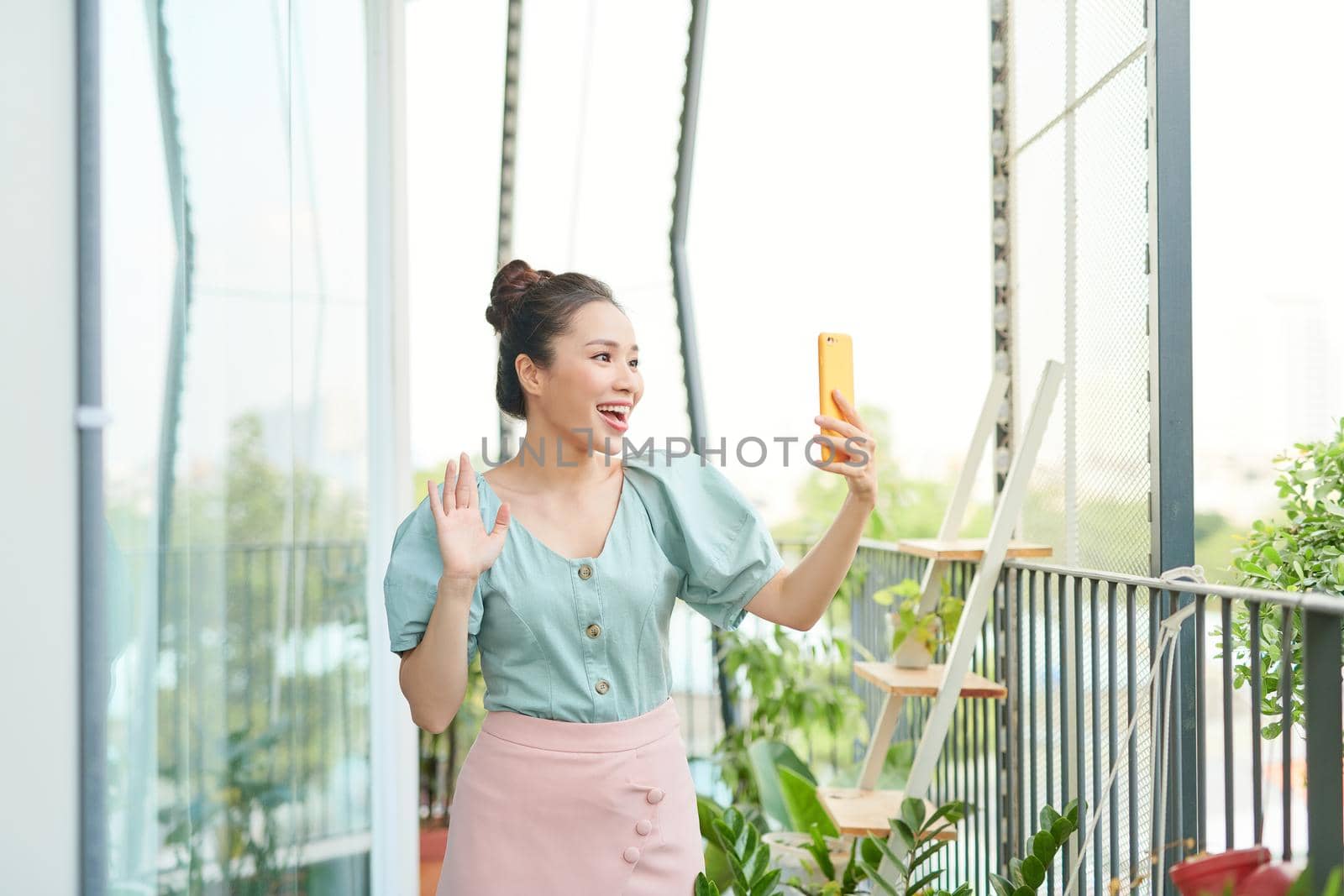 Cheerful young Asian girl having a video call with her phone when standing o balcony.