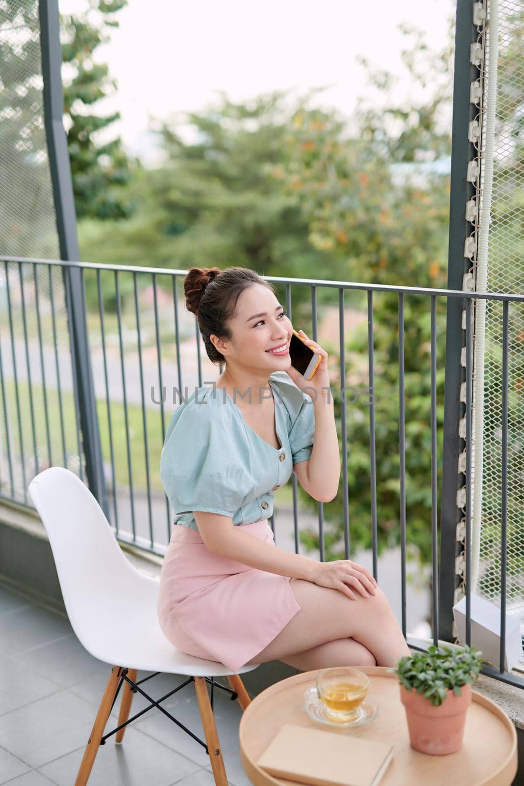 Asian young woman enjoying cup of coffee while using phone on the balcony.