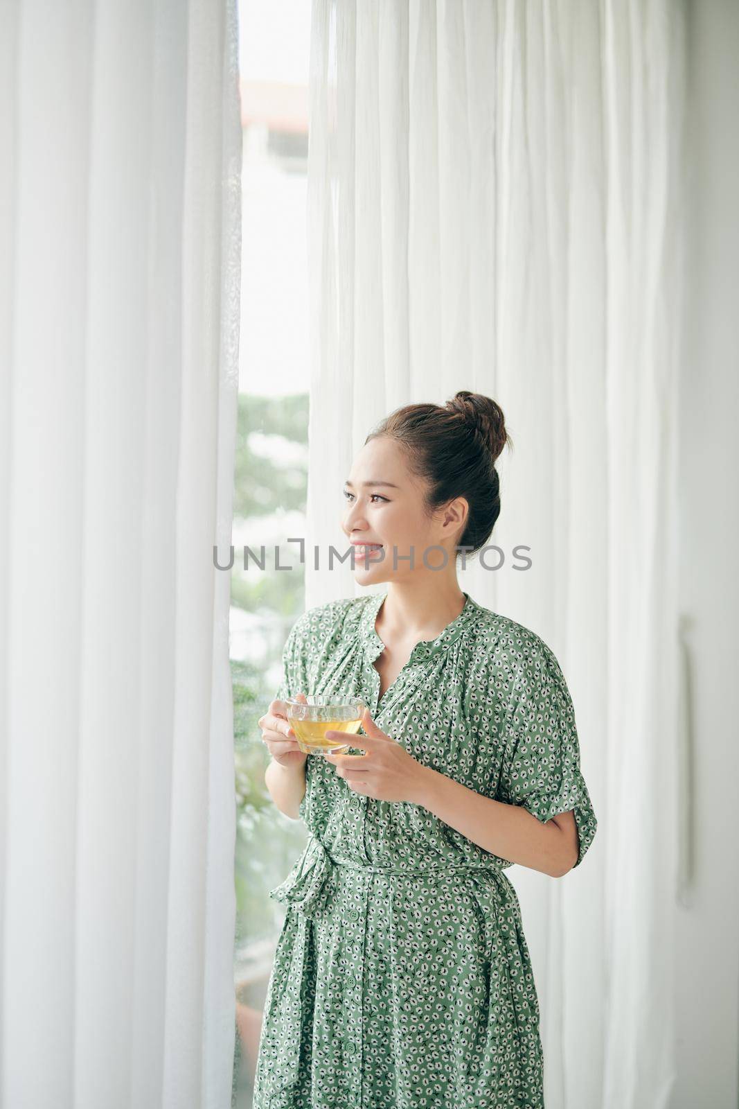 Beautiful Young Woman near big Window holding cup and relax inside home by makidotvn