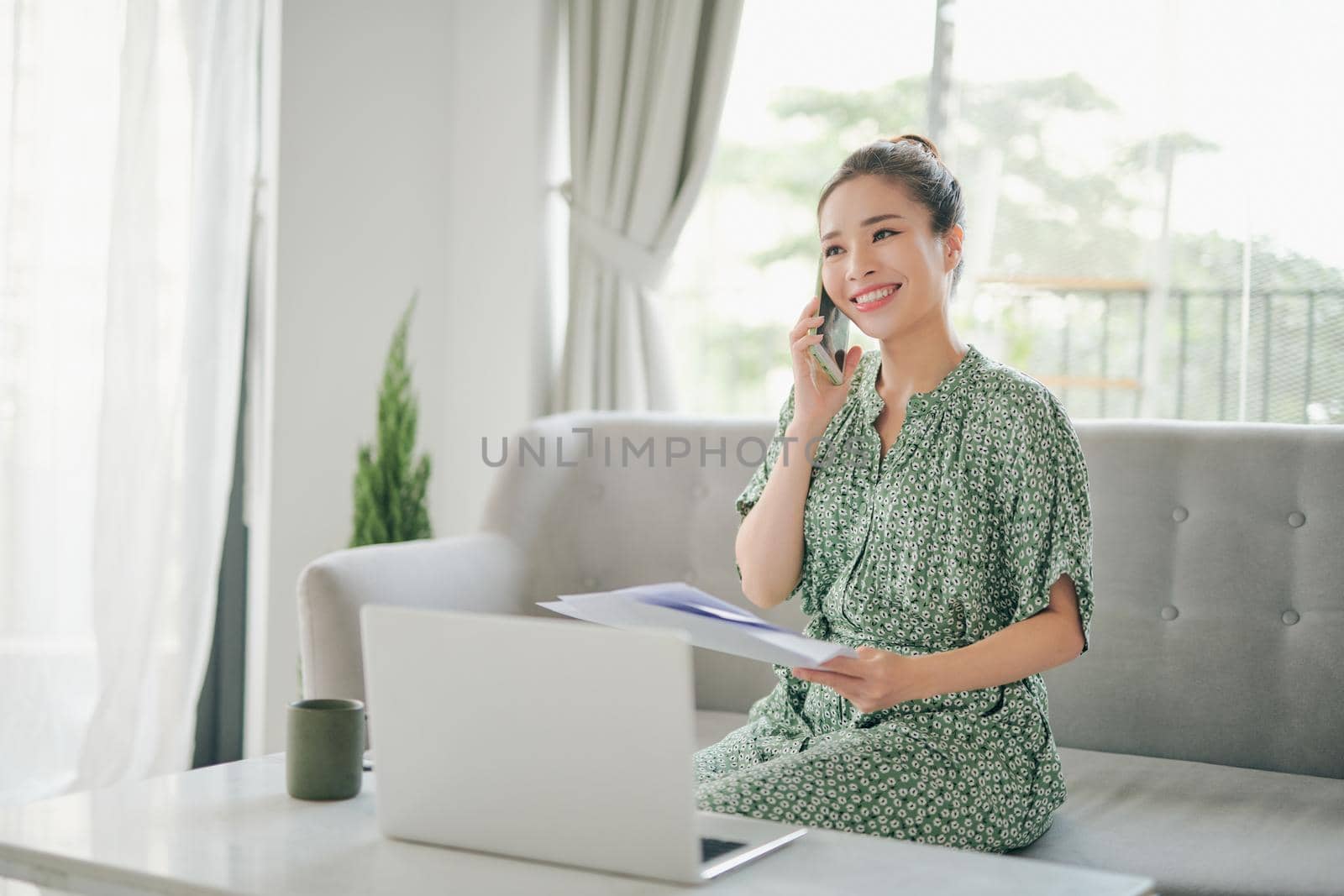 Happy young woman freelancer talking on mobile phone and using laptop, working from home during isolation, copy space. 