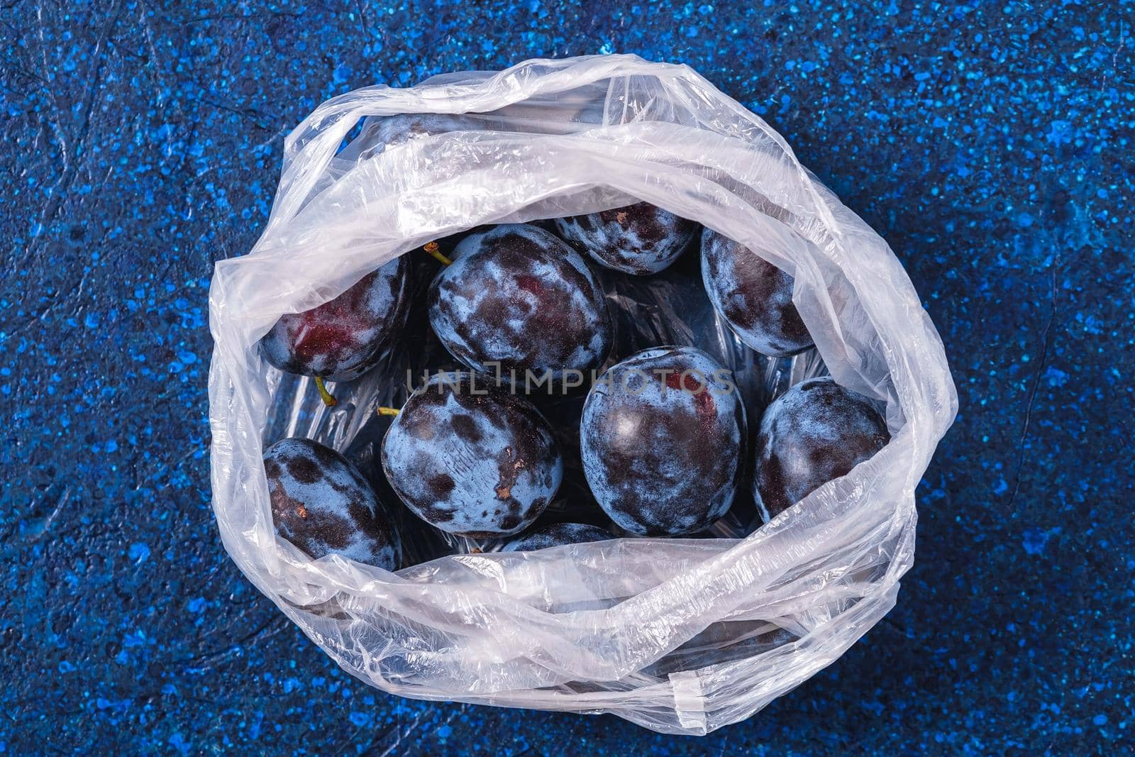 Fresh ripe plum fruits in plastic bag package on blue abstract background, top view
