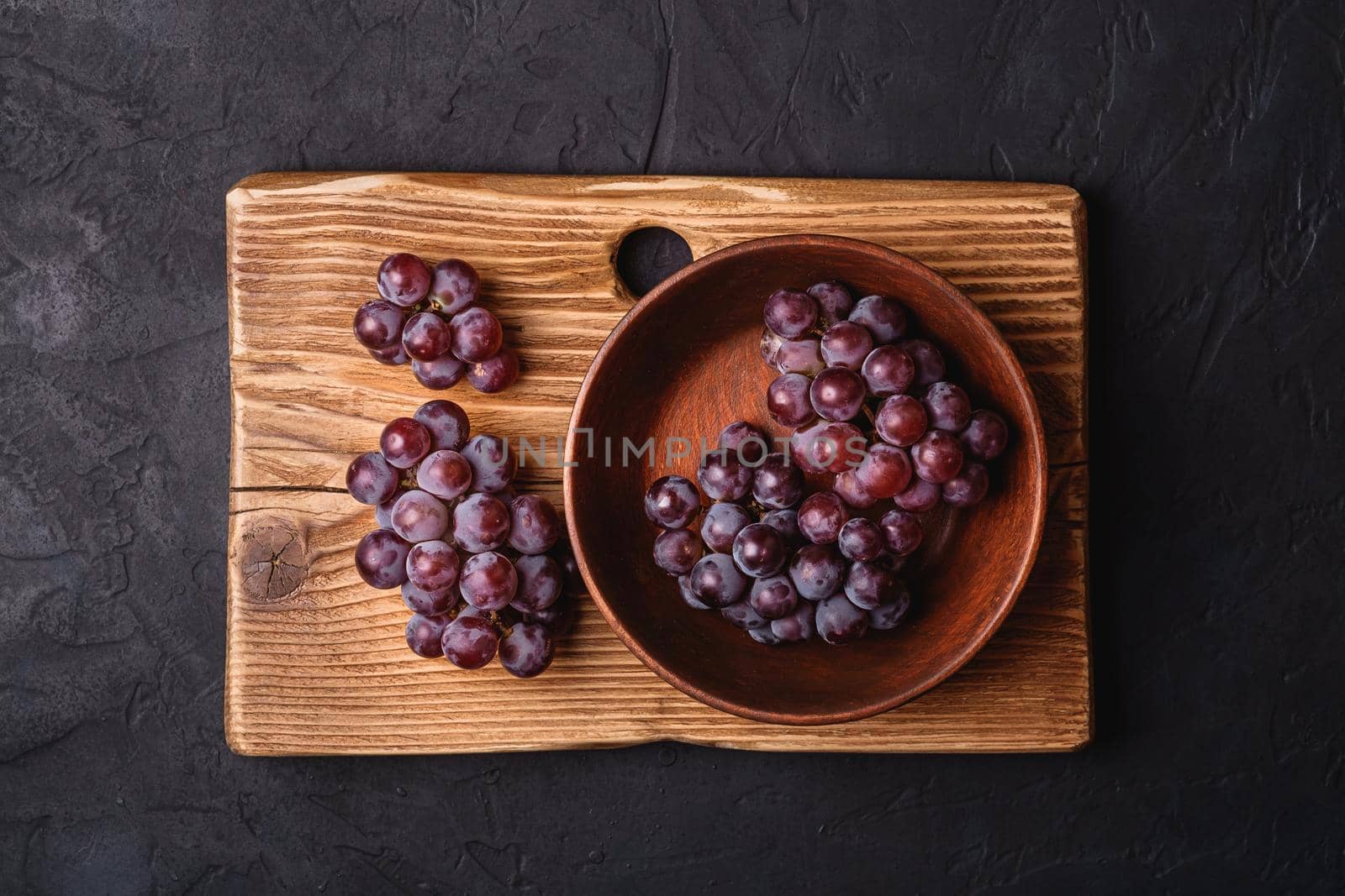 Fresh ripe grape berries in brown wooden bowl and cutting board on dark stone background, top view