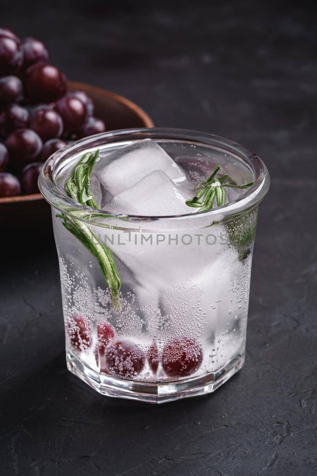 Fresh ice cold carbonated water in glass with rosemary leaf near to wooden bowl with grape berries, dark stone background, angle view by Frostroomhead
