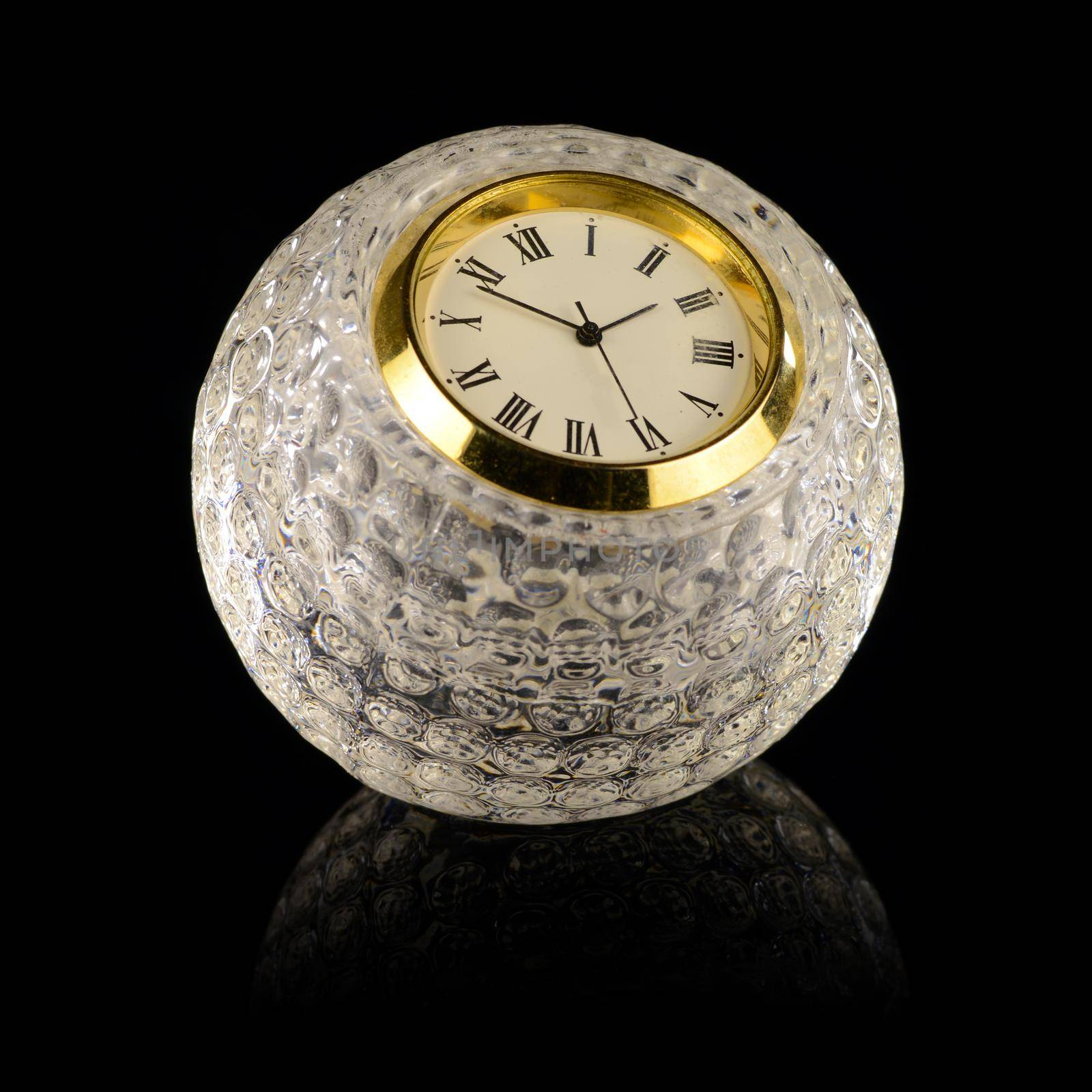 A crystal golf ball shaped clock with gold trim and almost striking three o'clock isolated over a pure black background.