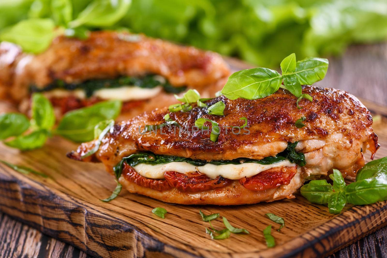 Baked chicken breast with mozzarella, sun-dried tomatoes, spinach. The meat is served on a wooden board with basil and lettuce.