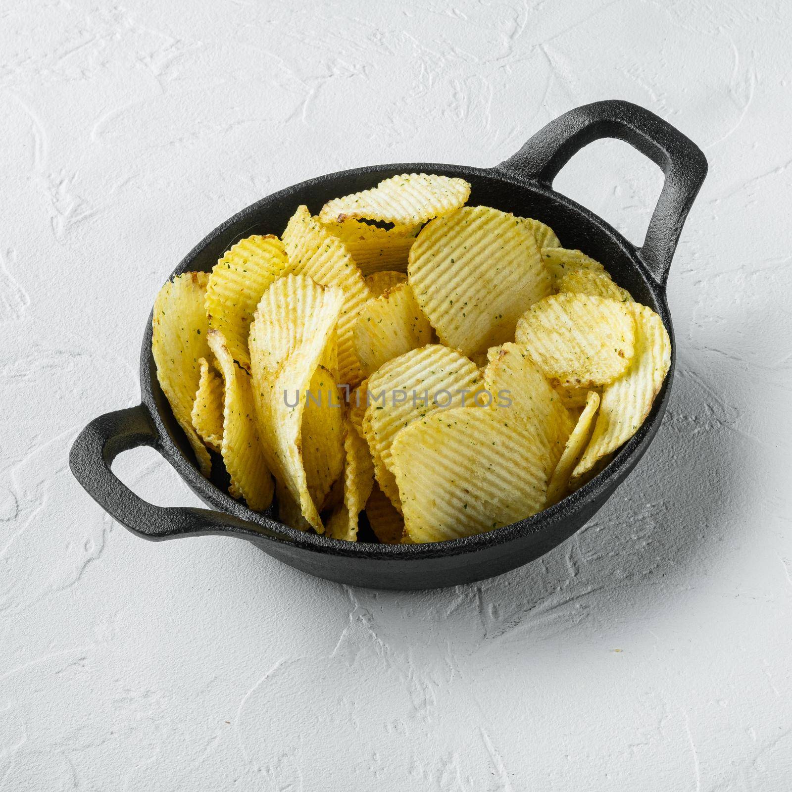 Crispy potato chips. Slices of potato, roasted with sea salt set, in cast iron frying pan, on white stone surface, square format