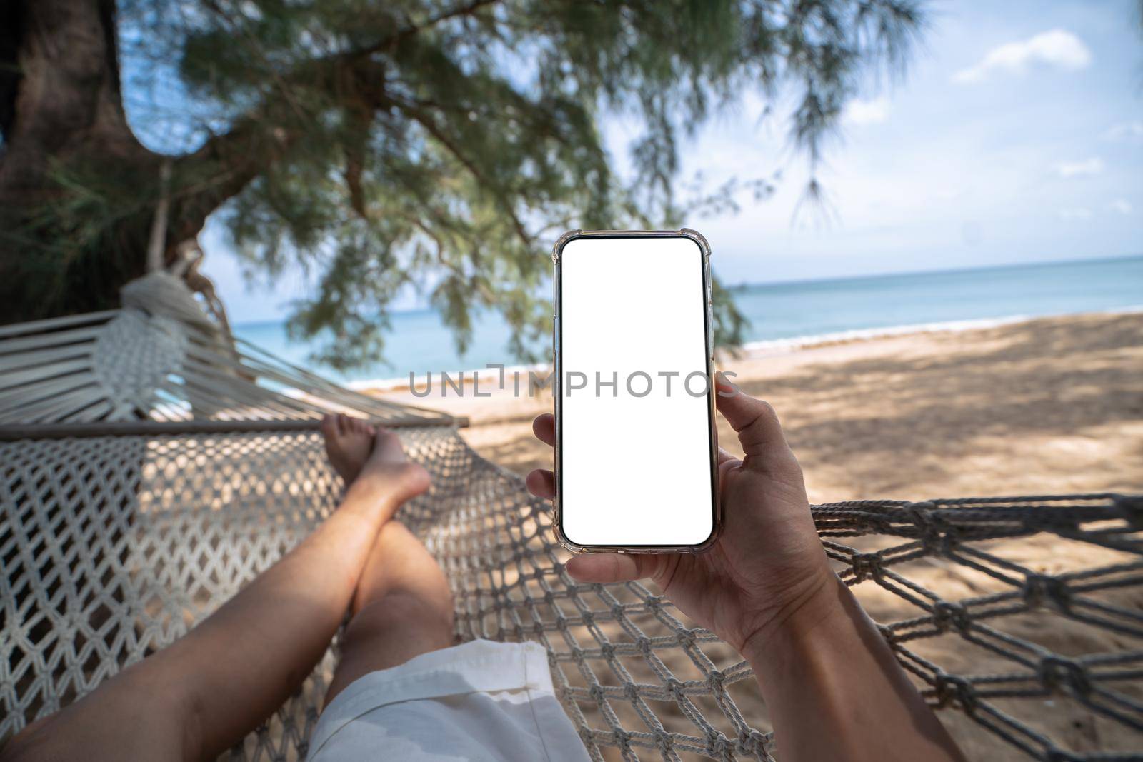 Hand holding mock up mobile with white sceen while laying on hammock swing between trees on the beach. by sirawit99