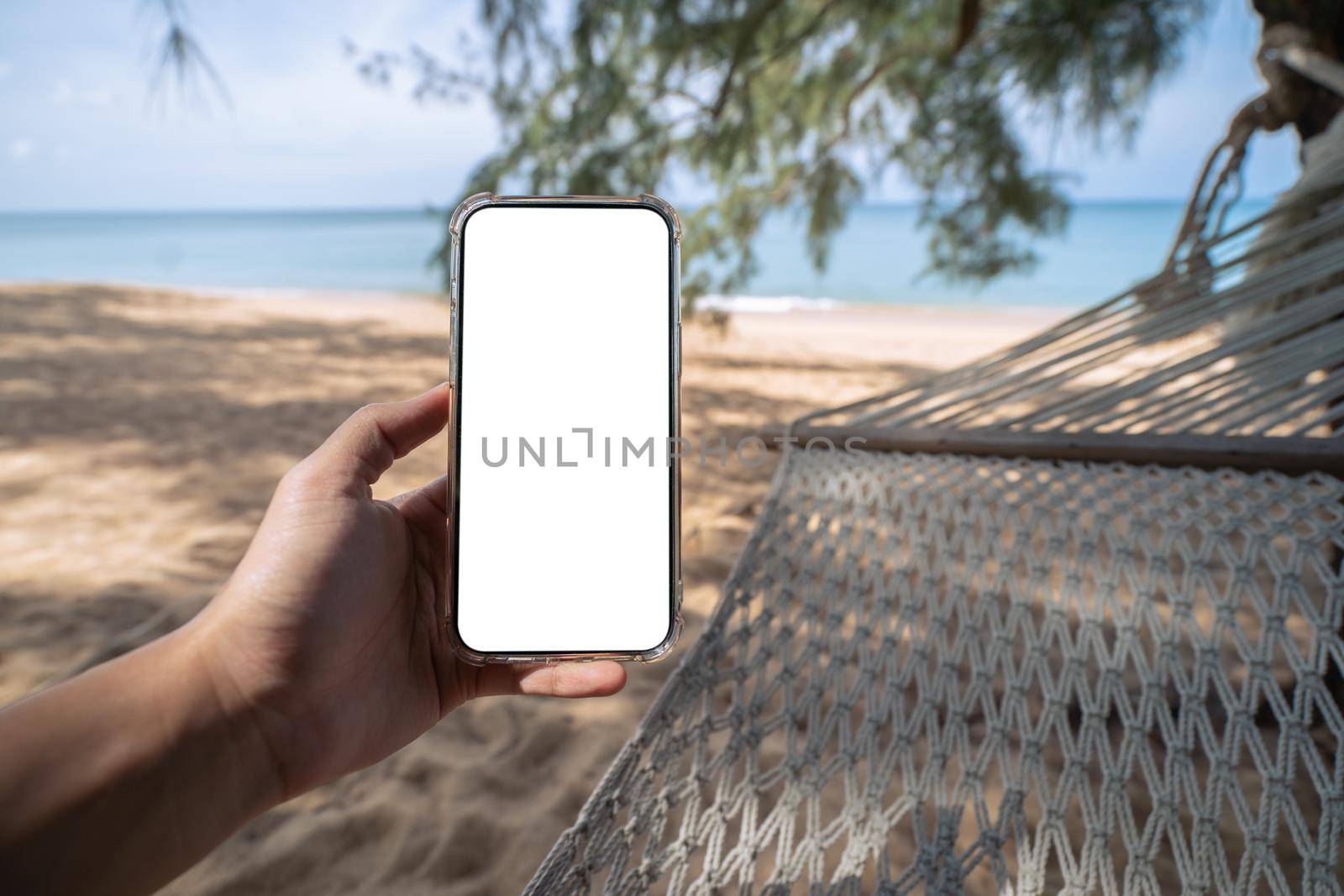 Hand holding mock up mobile with white sceen while laying on hammock swing between trees on the beach. by sirawit99