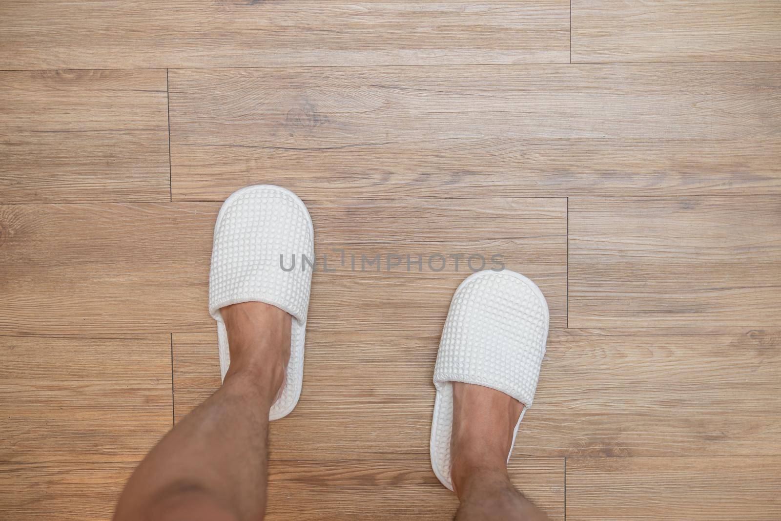 Slippers on man legs . Feet wearing white house slippers stand on brown wood floor.