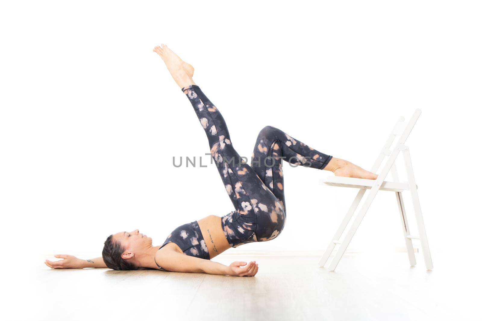 Young sporty attractive woman in bright white yoga studio, stretching and relaxing during restorative yoga using white chair as a gadget. Healthy active lifestyle.