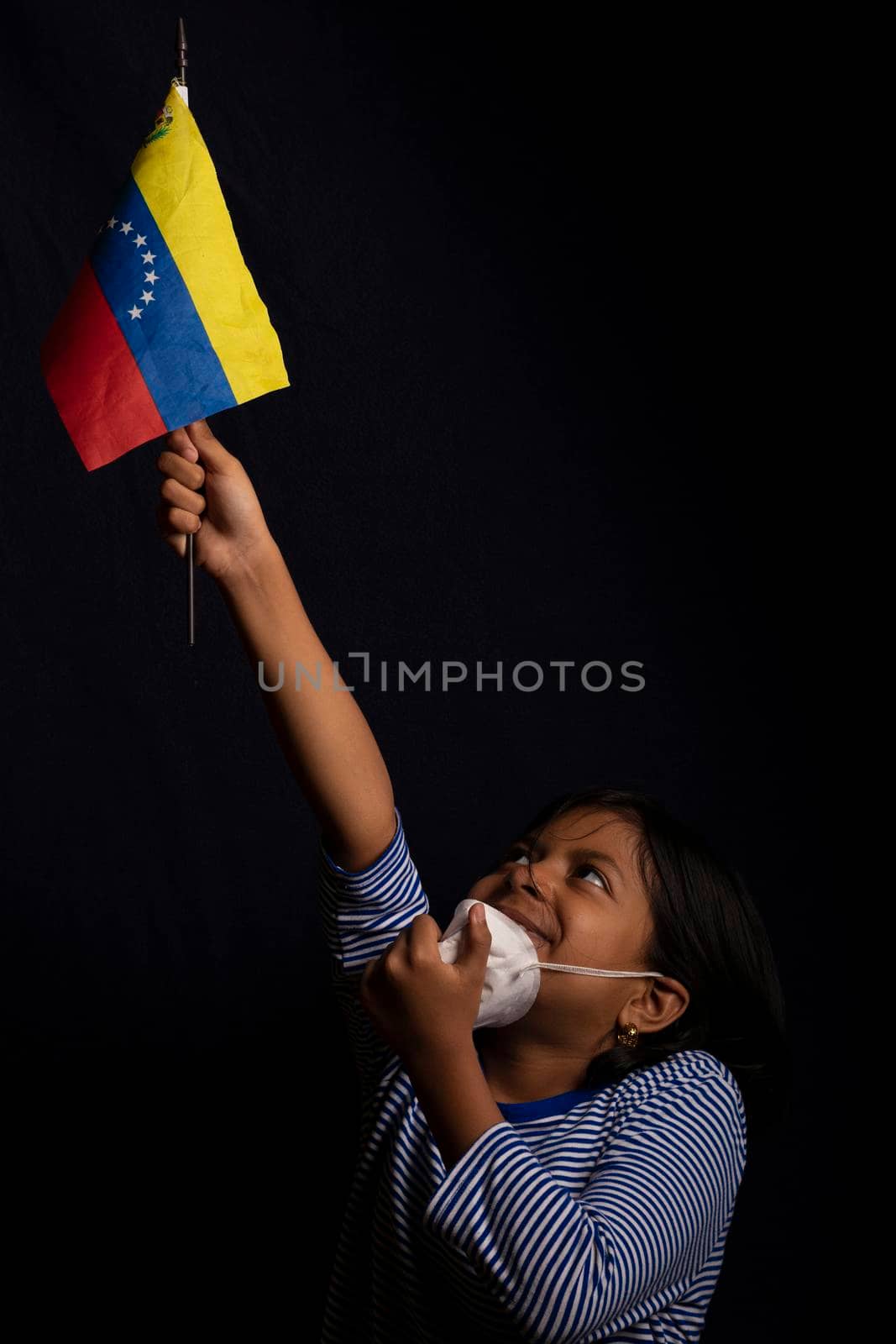 Portrait of little Venezuelan girl wearing medical mask and holding hopefully the flag of Venezuela
