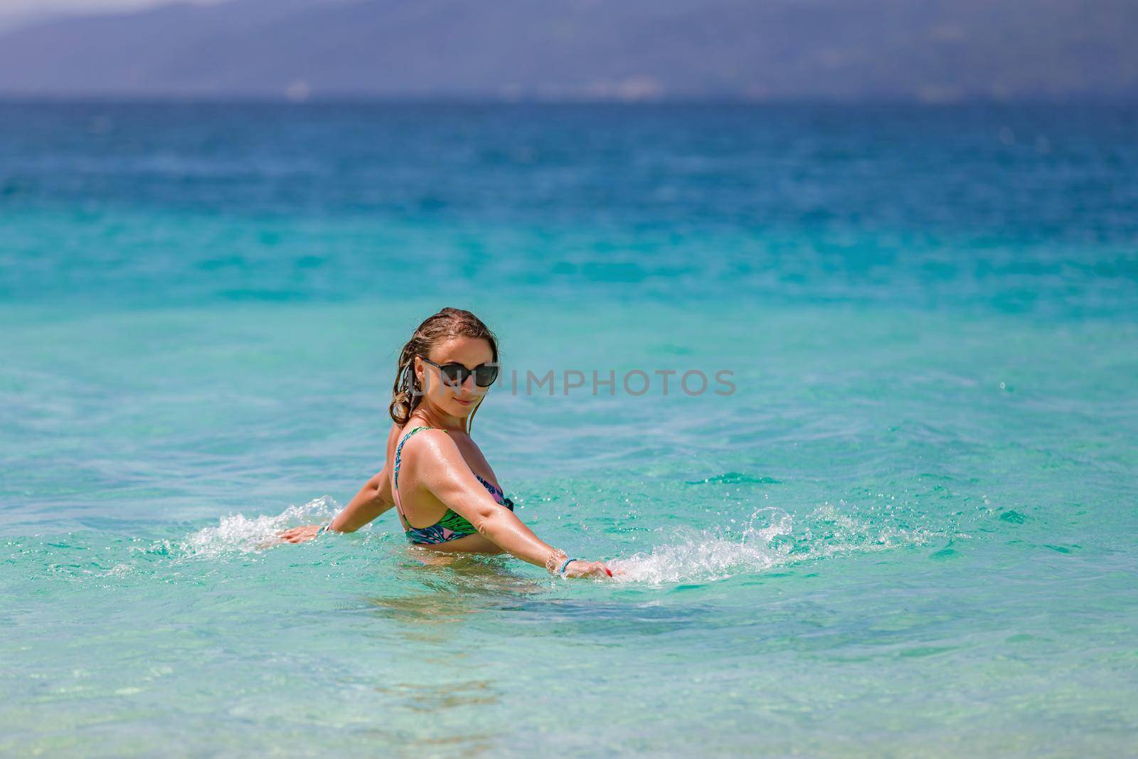 Young beautiful girl splashes in the blue water of the ocean. Rejoices and laughs on vacation