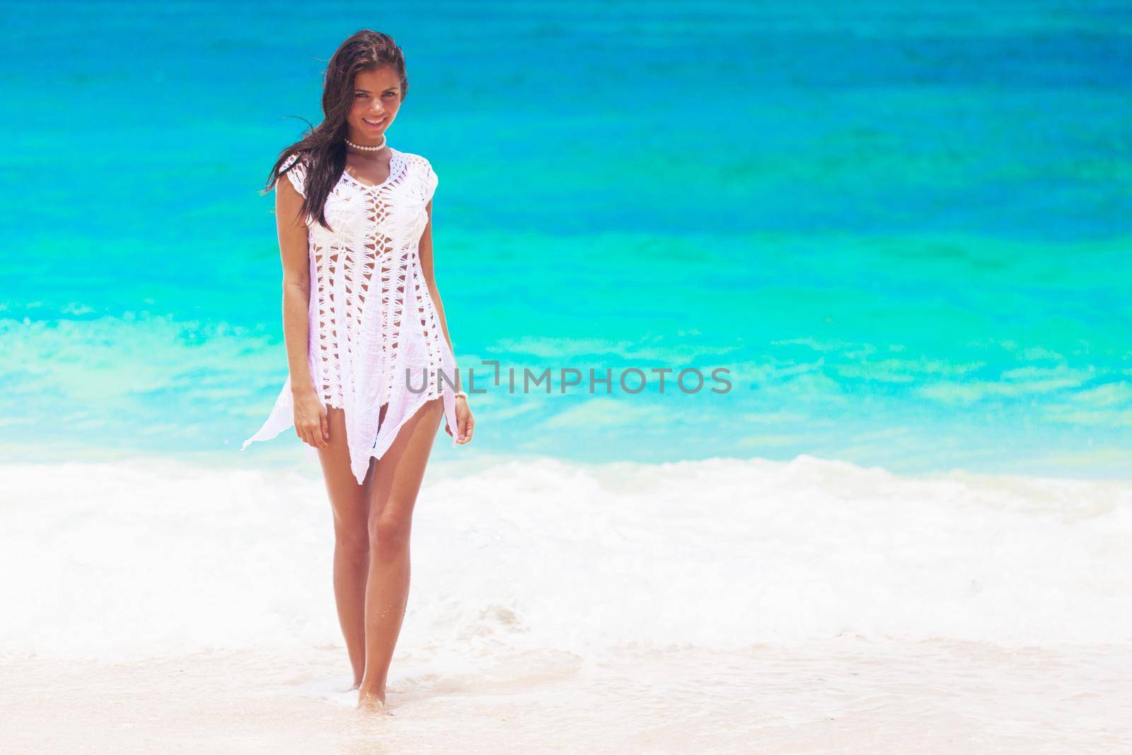 Happy young woman wearing white dress is walking on the beach of tropical sea and smiling