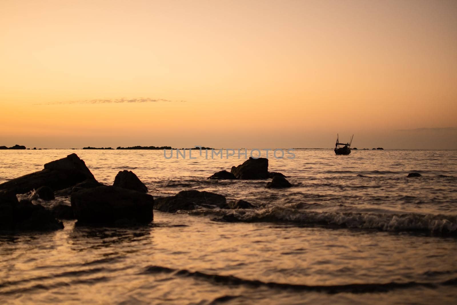 A bright golden orange sunset over a beach with rocks and a boat in the water waves near Ngwesaung beach, Irrawaddy, western Myanmar