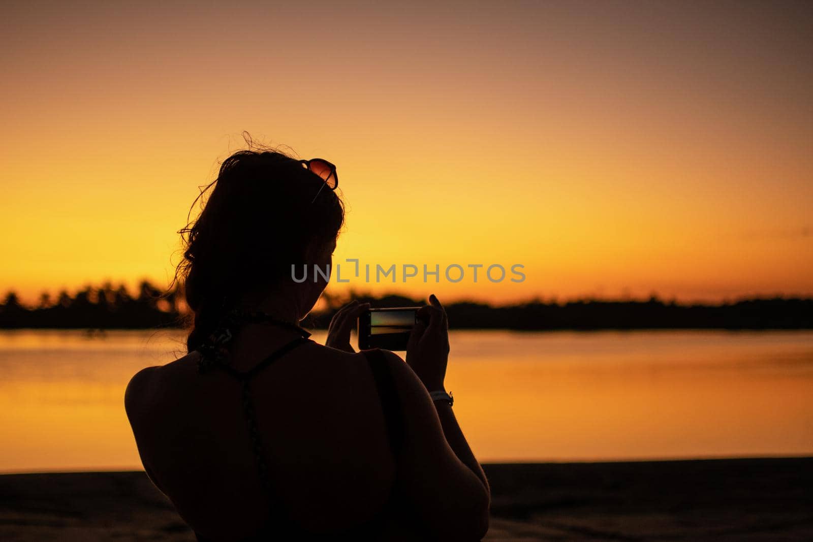 A woman with sunglasses takes a phone photo of the bright orange sunset and reflection near Ngwesaung, Irrawaddy, western Myanmar