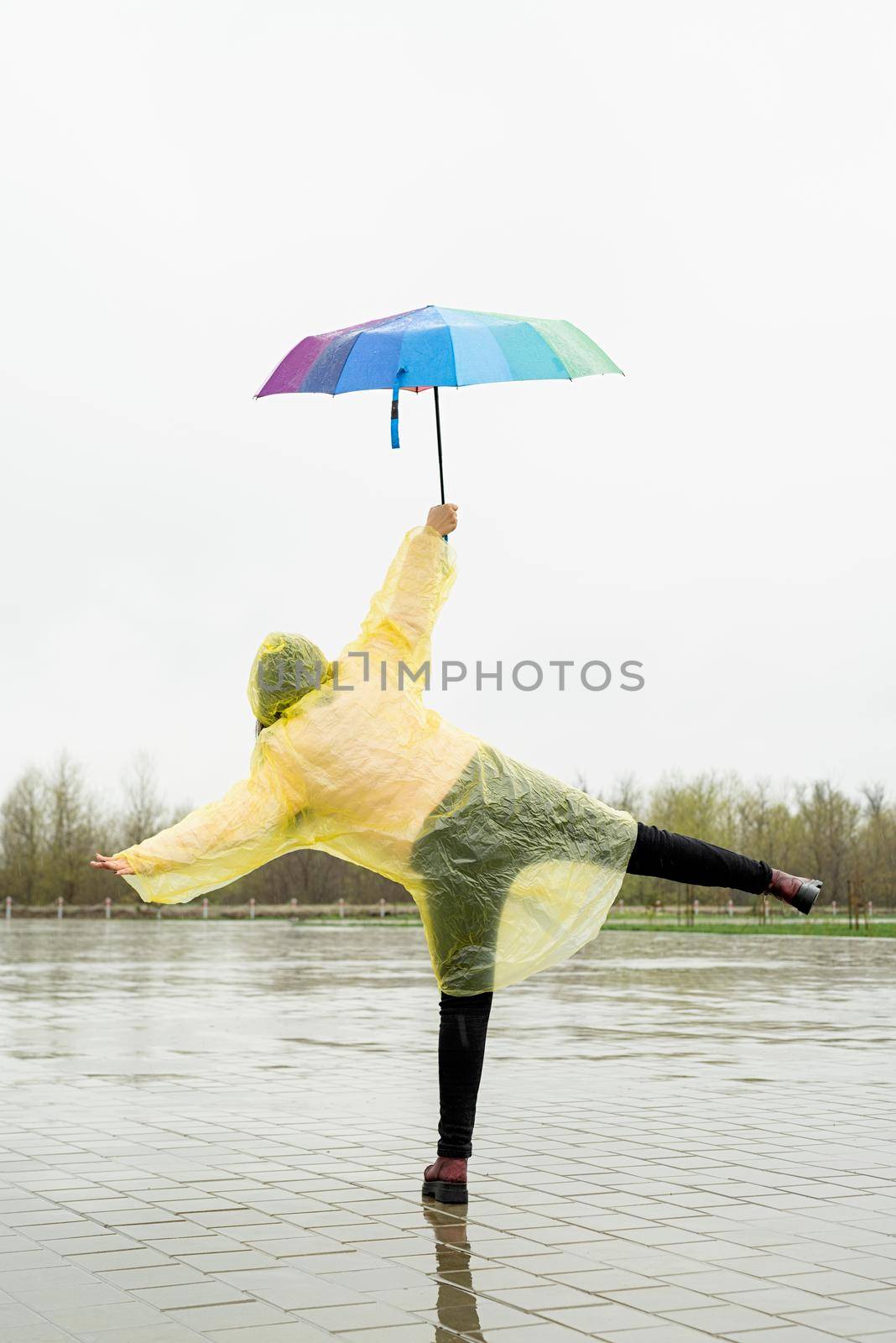 Beautiful brunnette woman in yellow raincoat holding rainbow umbrella dancing out in the rain