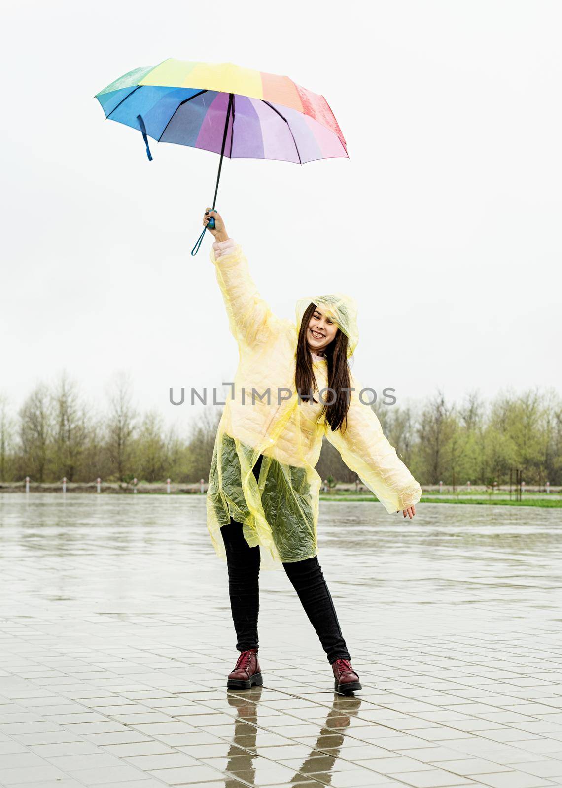 Beautiful brunette woman in yellow raincoat holding rainbow umbrella out in the rain
